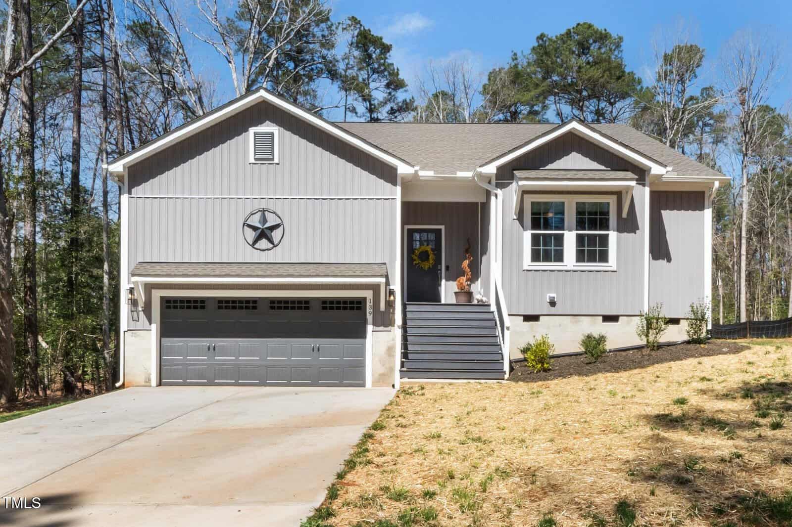 A modern gray house with a two-car garage, front steps leading to a yellow front door with a decorative wreath, a star mounted above the garage, and a small landscaped yard. Tall trees are visible in the background.