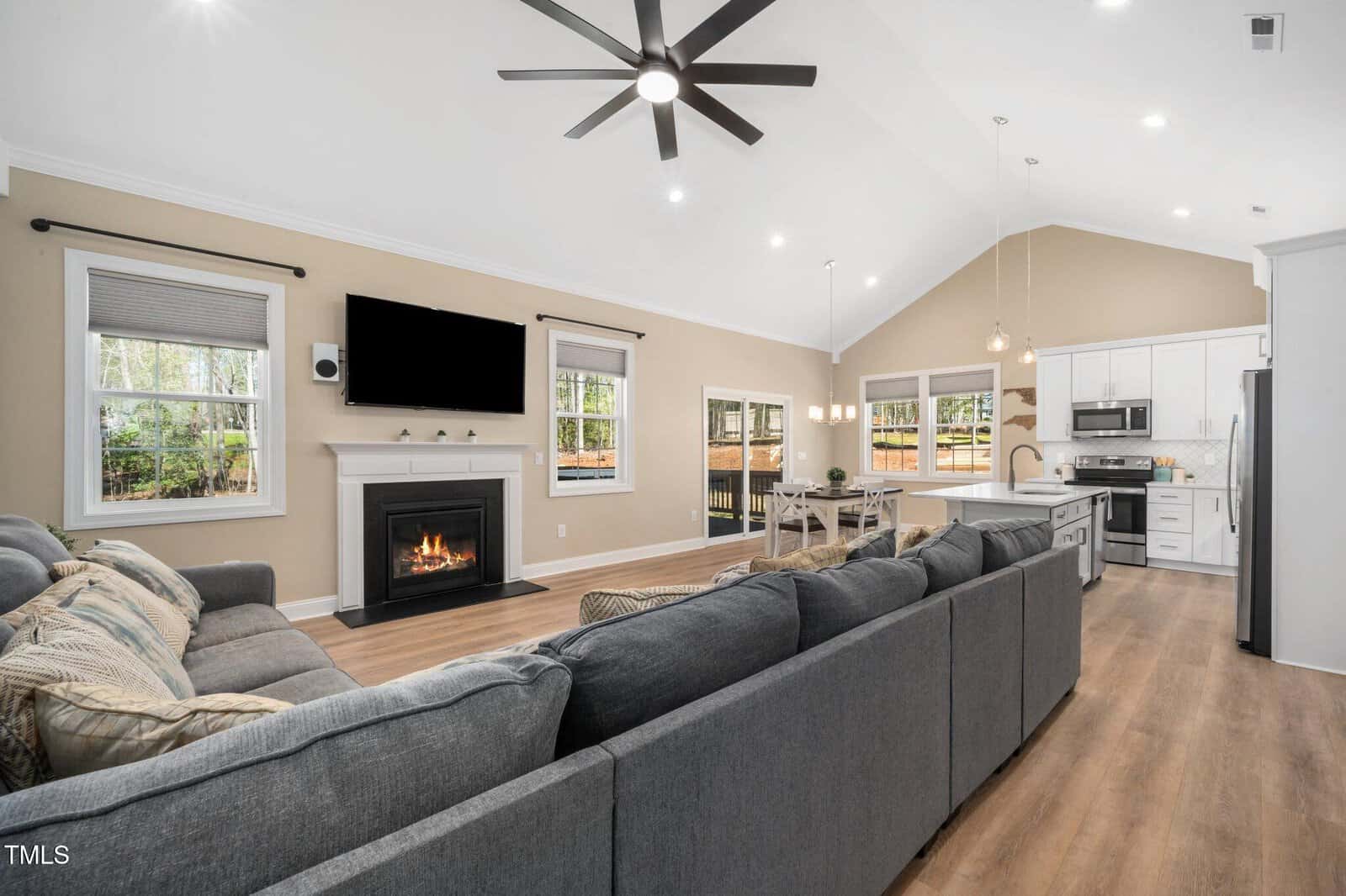 Open-concept living room and kitchen with a gray sectional sofa, fireplace under a wall-mounted TV, wooden flooring, dining table, modern light fixtures, large windows, and neutral-colored walls.