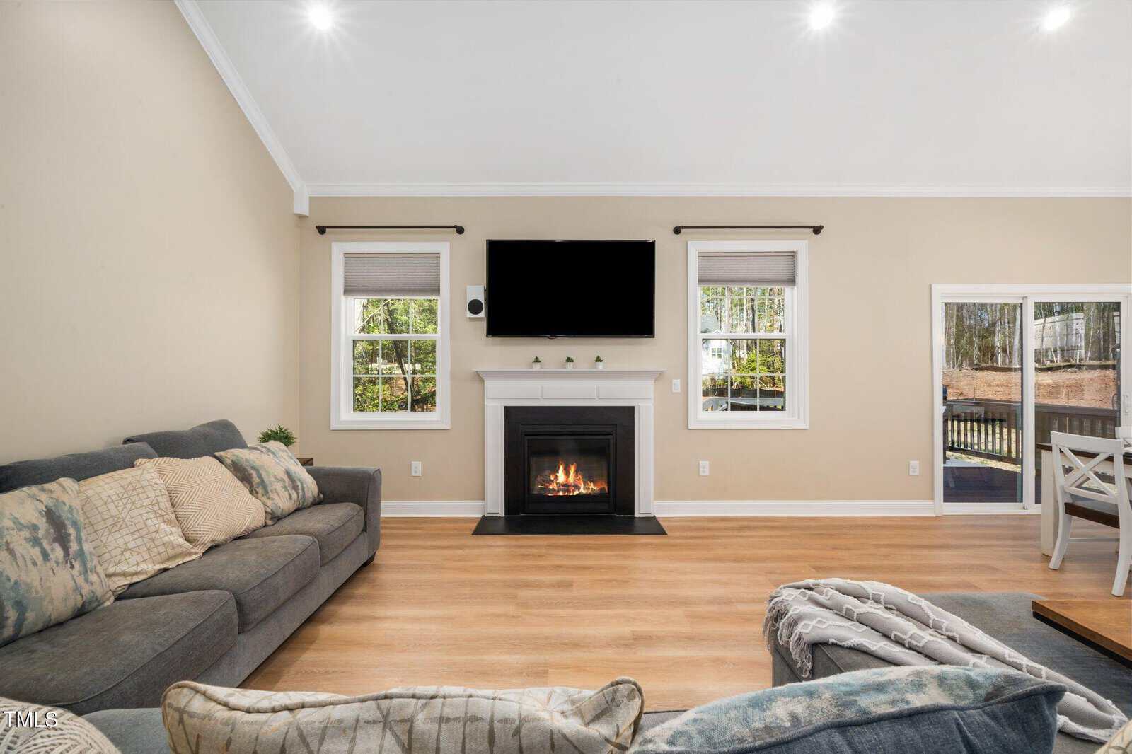 Living room with a gray sofa, armchair, fireplace under a wall-mounted TV, hardwood floors, beige walls, two windows, glass door to deck, and a small dining area visible on the right.