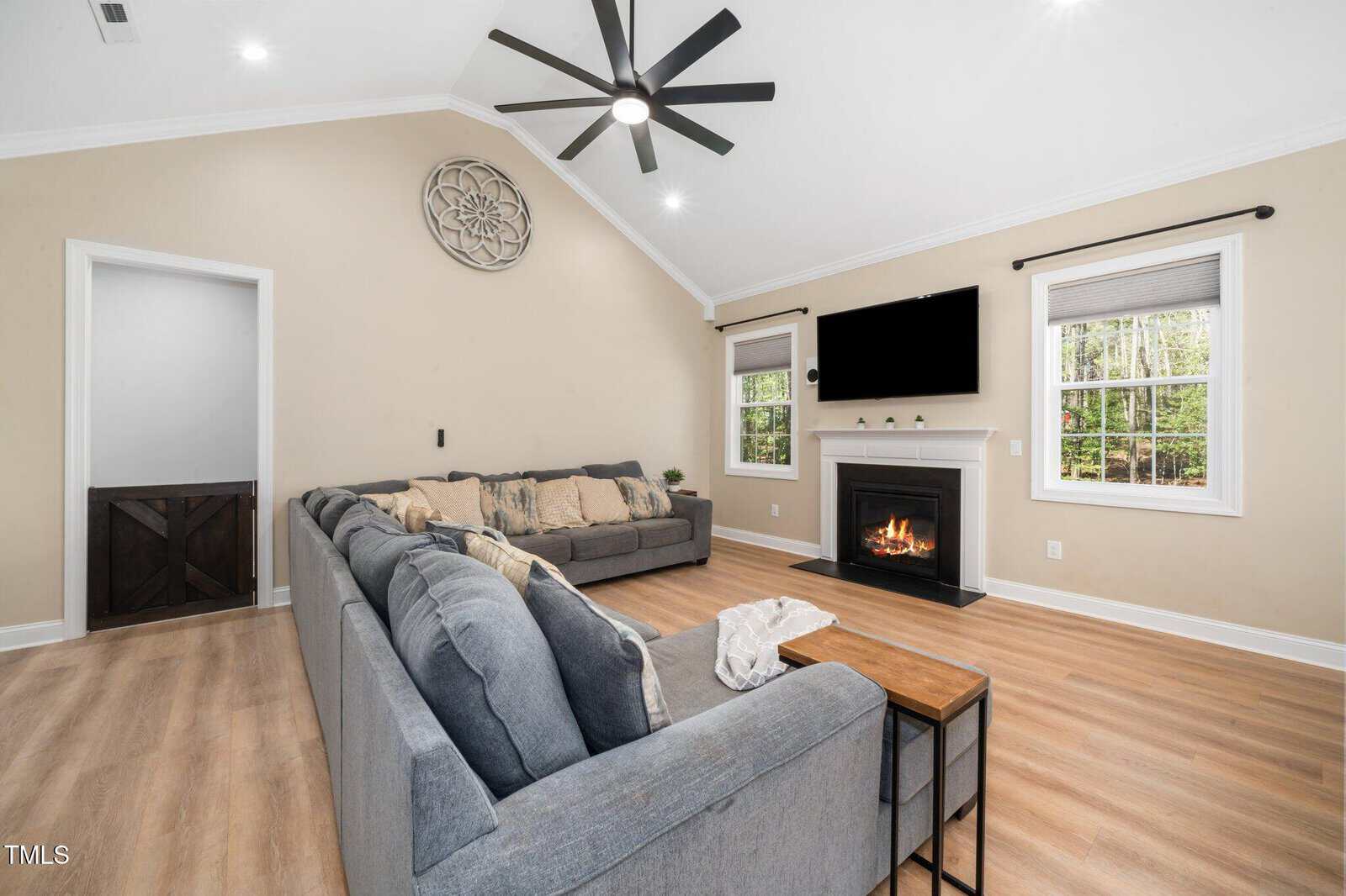 Living room with a gray sectional sofa, wood flooring, a fireplace with a TV above, two windows, beige walls, a ceiling fan, and wall decor. A small table is beside the sofa, and trees are visible outside.