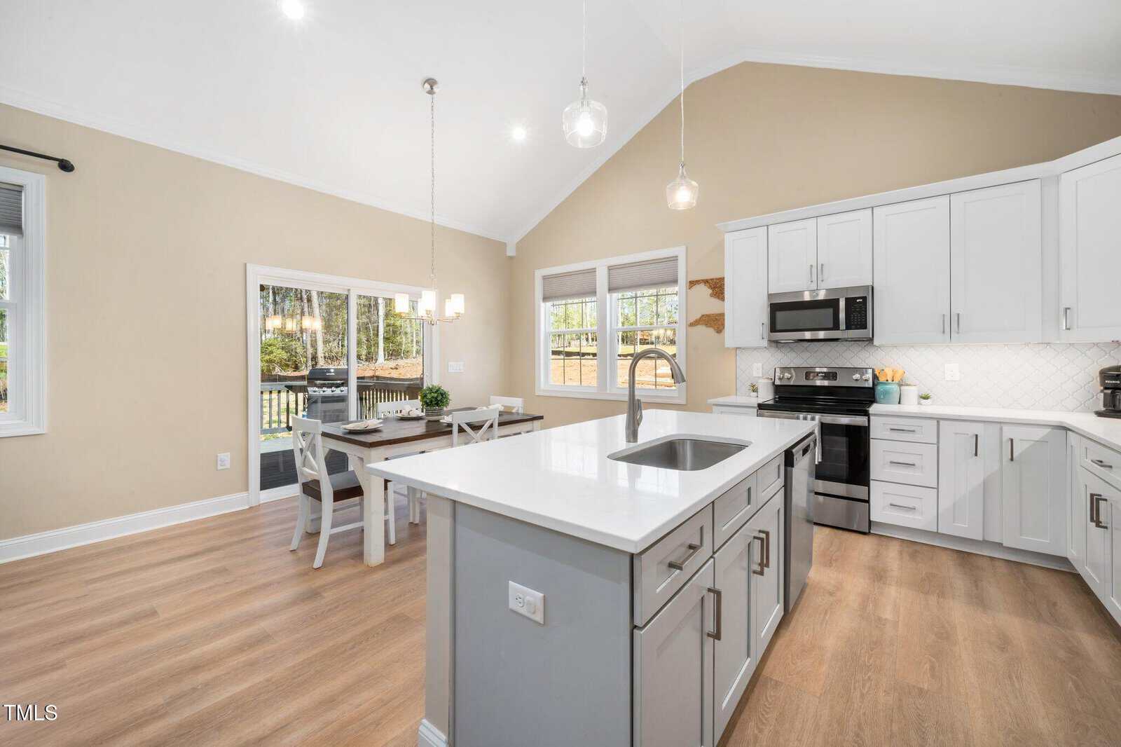 Modern kitchen with white cabinets, stainless steel appliances, an island with sink, and pendant lights. There is a dining table near large windows with views of a fenced backyard. Light wood flooring throughout the space.