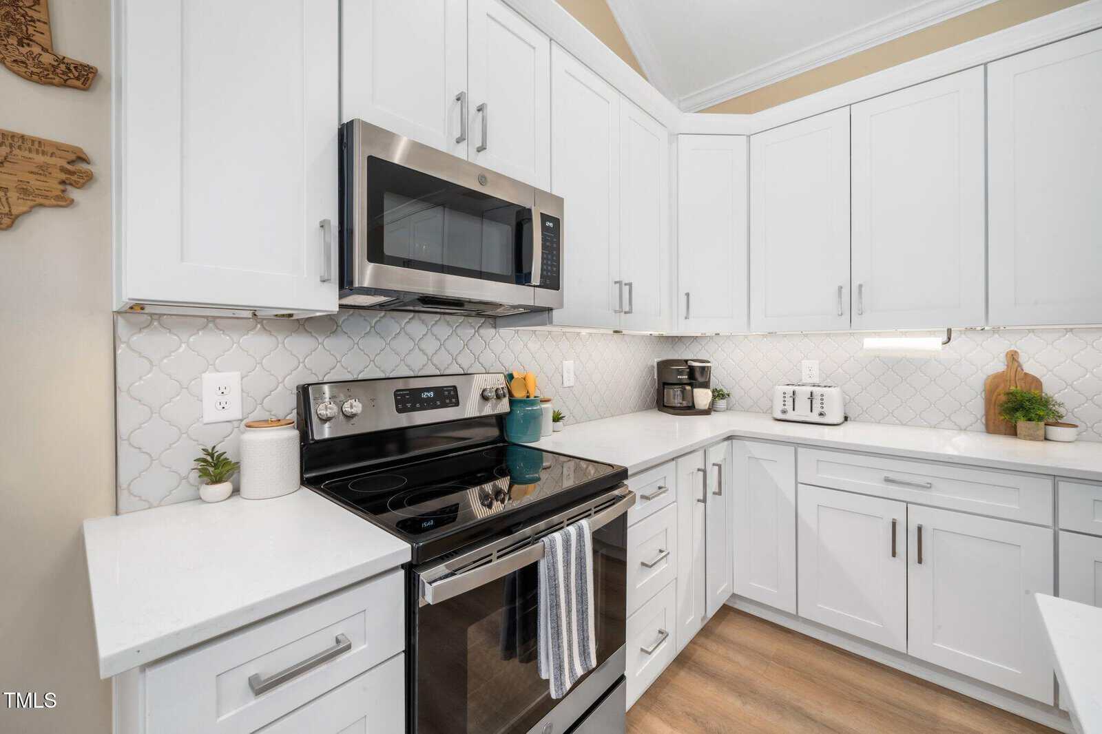 Modern kitchen with white cabinets, stainless steel appliances, electric stove, microwave, toaster, coffee maker, and small potted plants on white countertops. Light wood flooring and white tile backsplash are visible.