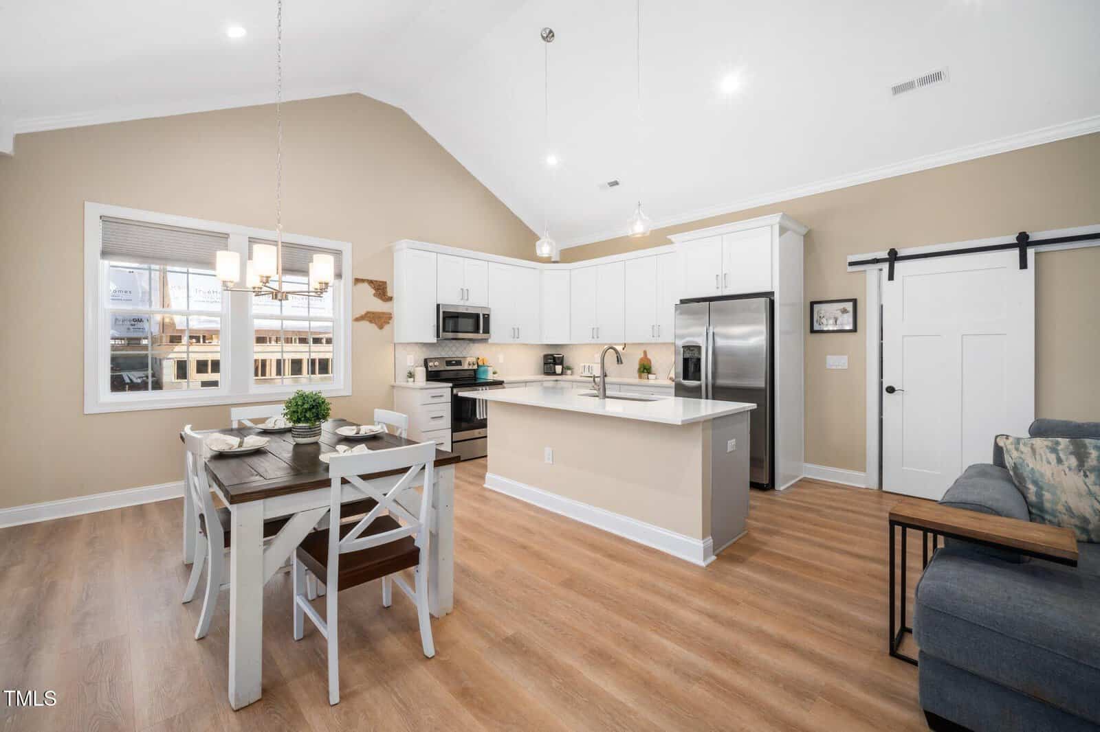 A modern kitchen and dining area with white cabinets, stainless steel appliances, a wooden dining table set for four, light wood flooring, and neutral-colored walls under a vaulted ceiling with recessed lighting.