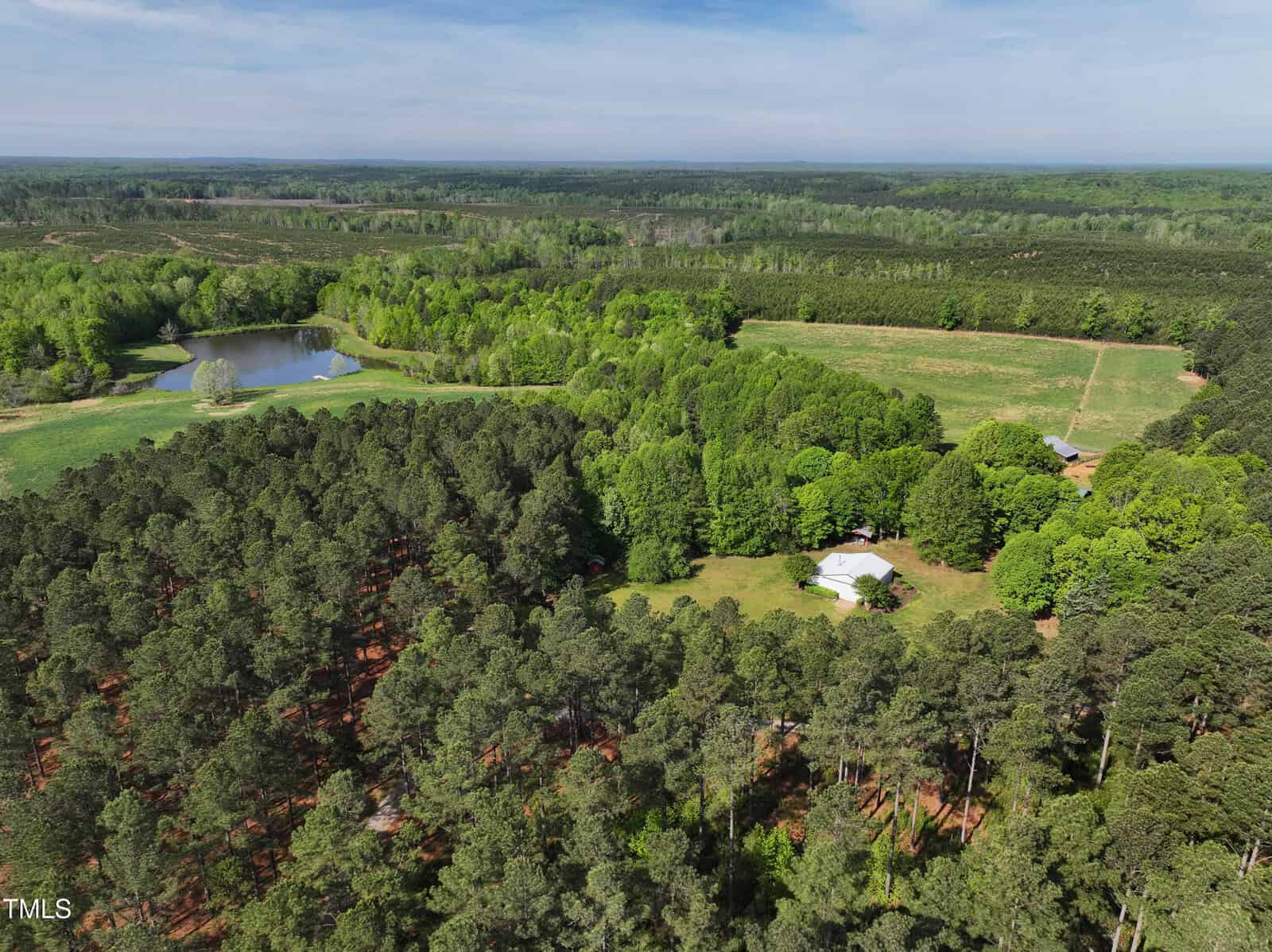 Aerial view of a rural property with a white house surrounded by trees, open fields, and a pond. Dense forest covers much of the landscape under a clear blue sky.
