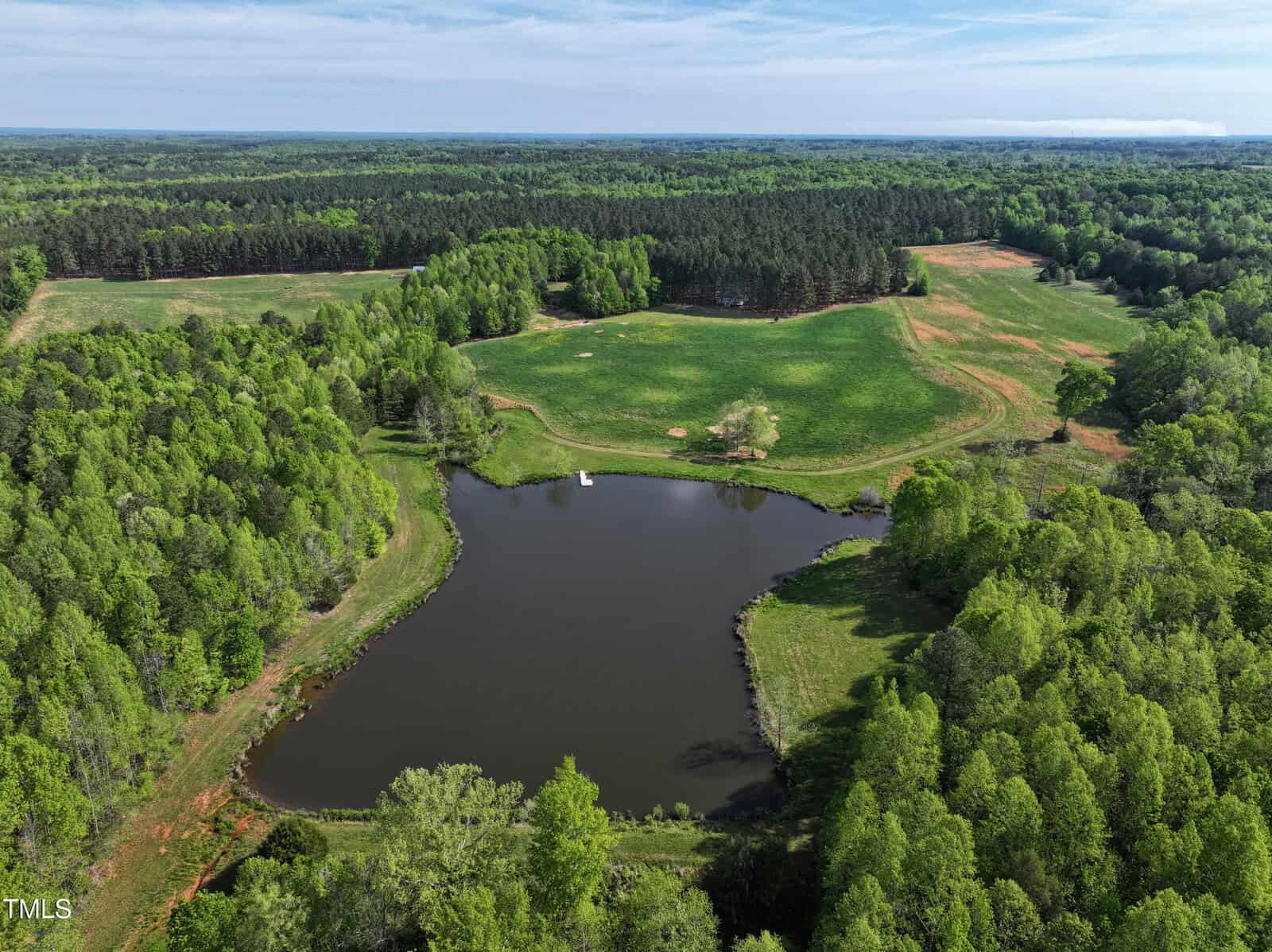 Aerial view of a rural landscape featuring a pond surrounded by trees, open grassy fields, and wooded areas under a partly cloudy sky. A small white object floats on the pond near the center.