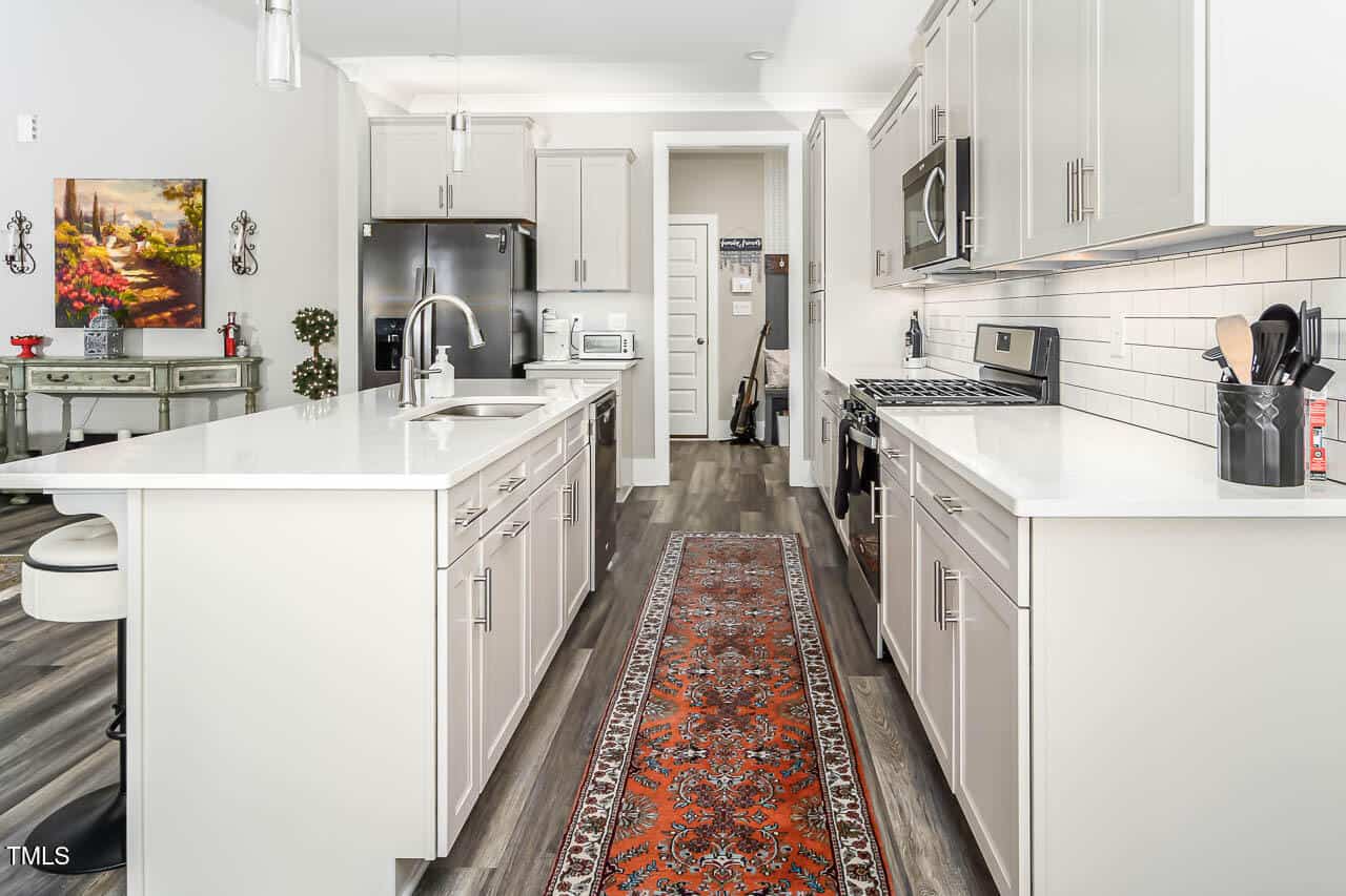 Modern kitchen with white cabinets, stainless steel appliances, a red patterned runner rug, and a white island. Black refrigerator and stove are on opposite sides. Wall art and kitchen utensils are visible in the background.