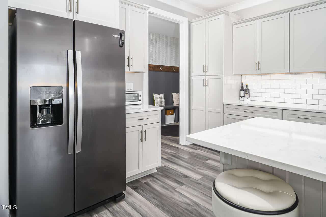 A modern kitchen with gray cabinets, stainless steel side-by-side refrigerator, white subway tile backsplash, marble countertop island, and wood-patterned flooring. A small countertop appliance and two bottles are visible.