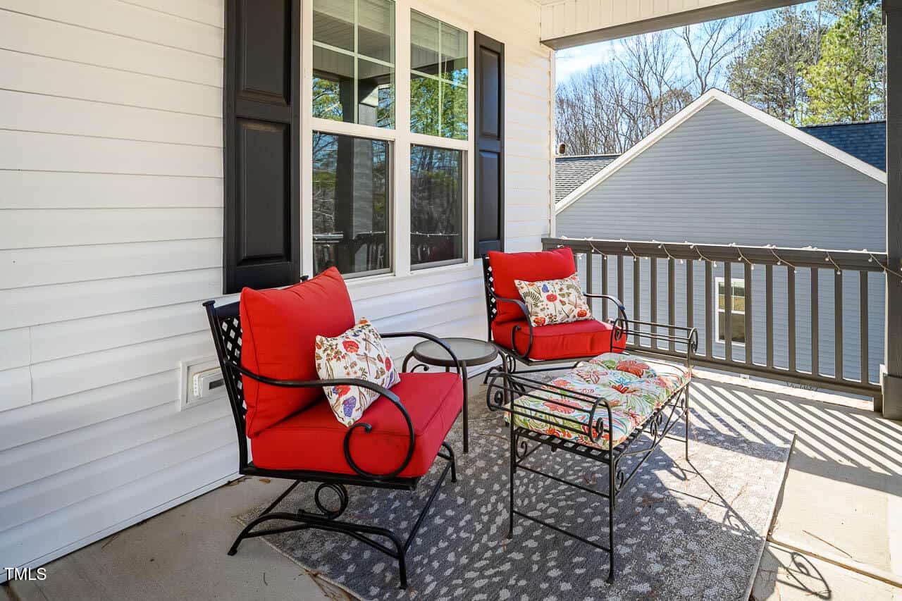 A covered porch with two black metal chairs with red cushions, floral accent pillows, a small round side table between them, and a neutral patterned outdoor rug; house siding and railing are visible.
