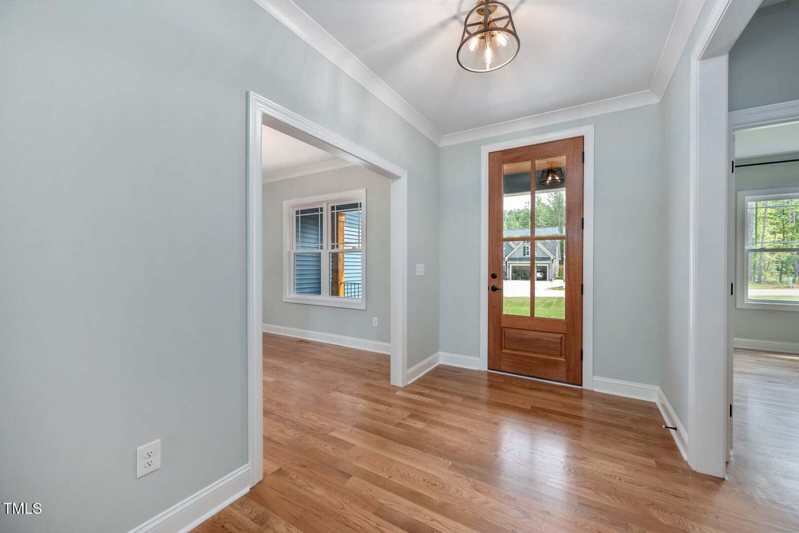 A hallway with light gray walls, wooden floors, and a wooden front door with glass panels. There are two open doorways leading to adjacent rooms with windows. Ceiling light fixture is visible.