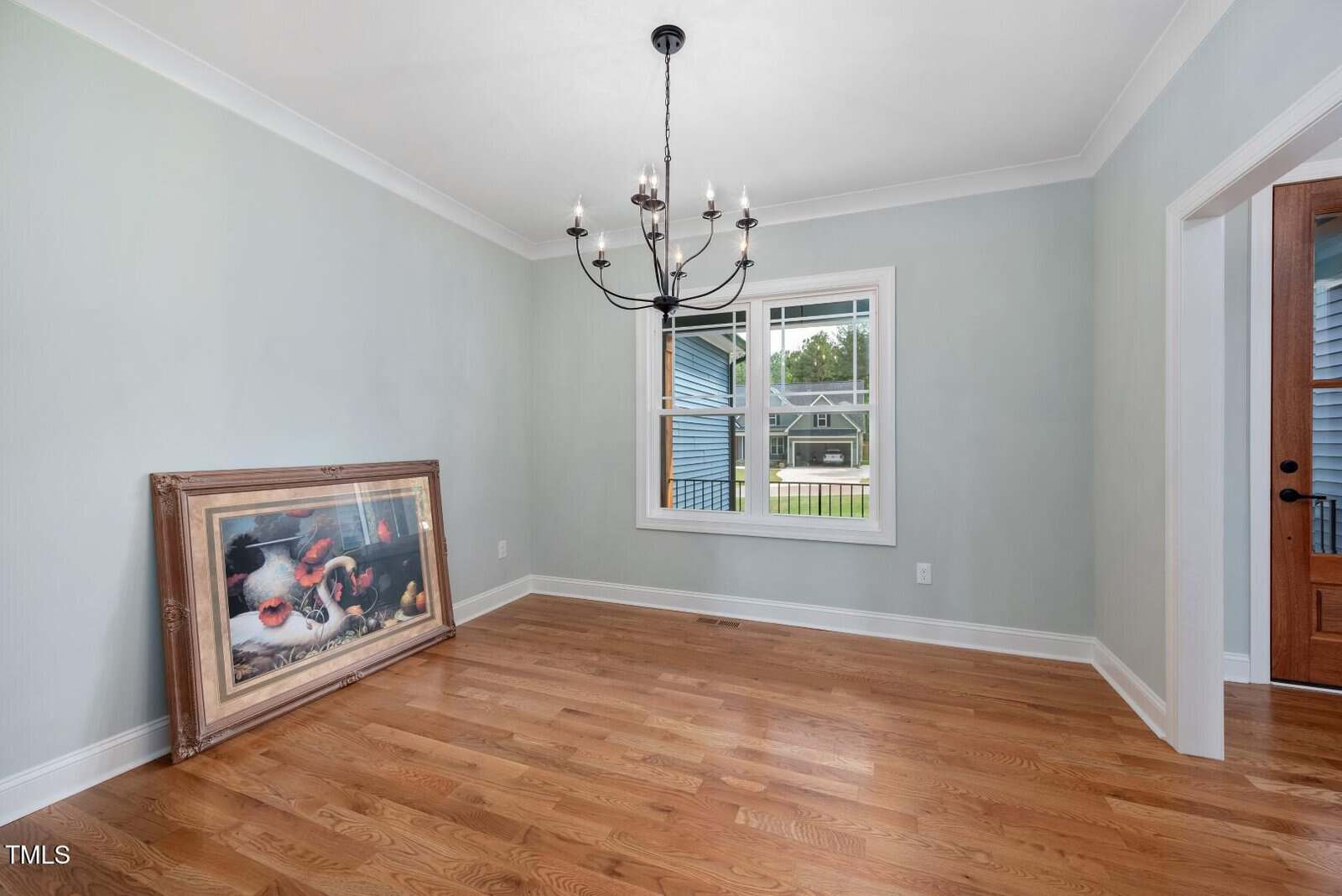 Unfurnished room with light blue walls, hardwood floors, a central black chandelier, a window, and a wooden door. A large framed picture leans against the wall.