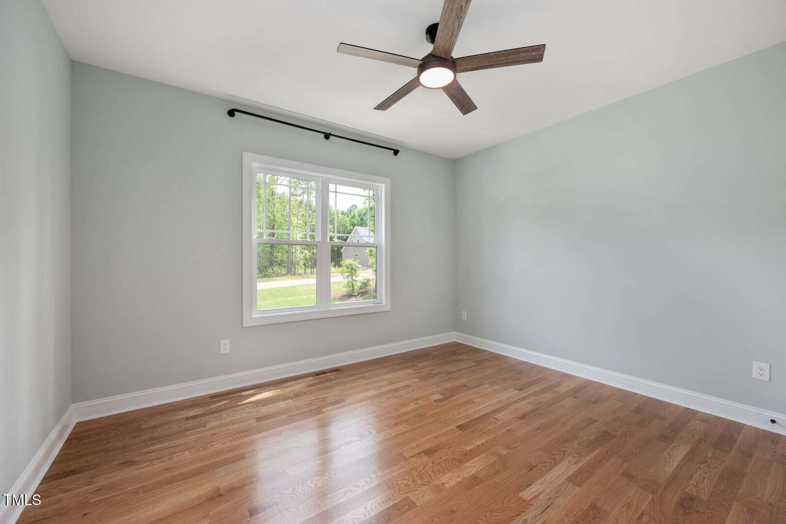 Empty room with light gray walls, hardwood floor, a ceiling fan, and a large window showing trees and grass outside. The space is well-lit by natural light from the window.