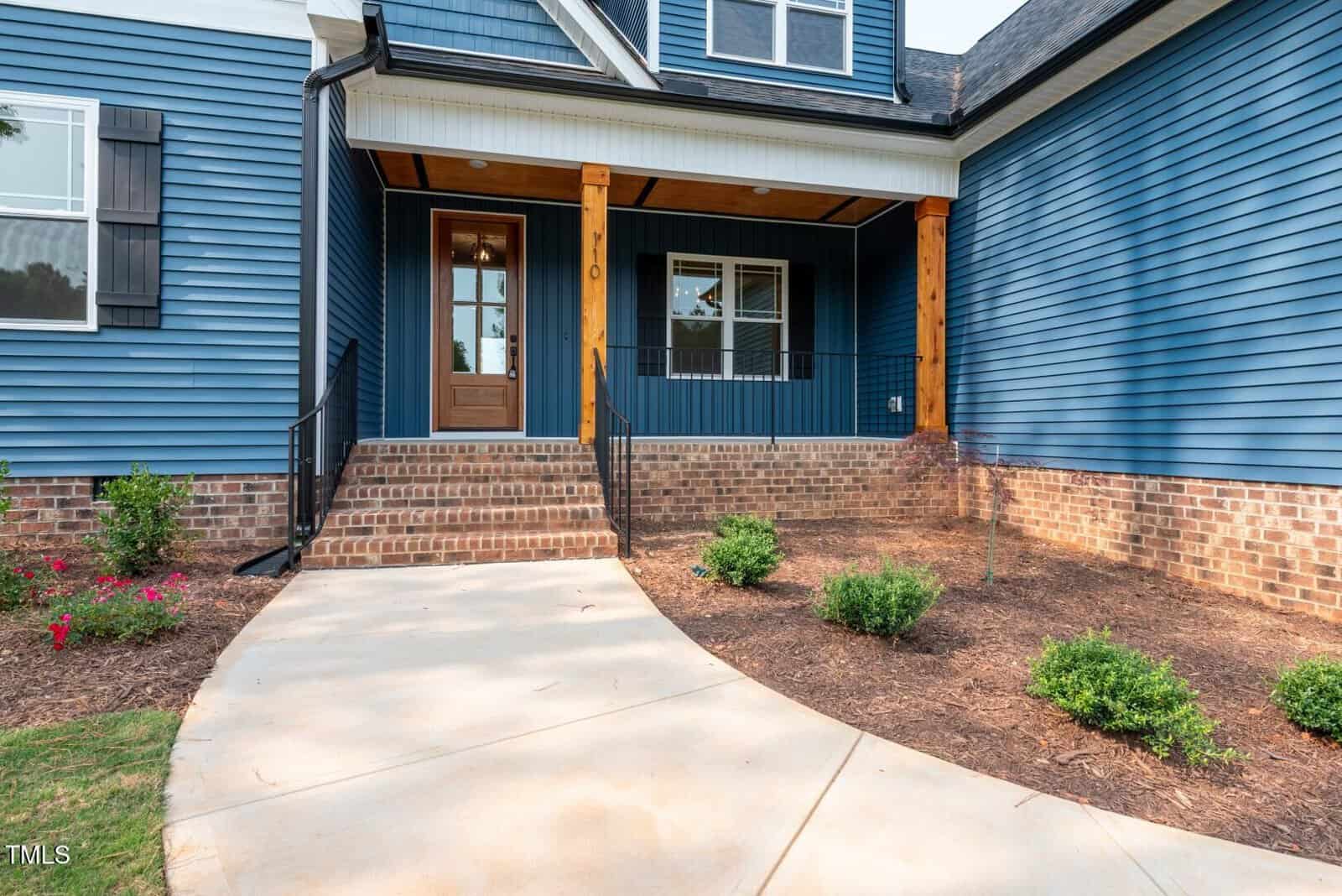 A blue house with brick accents features a covered front porch, wooden columns, black metal railing, and steps leading to a wooden front door. The landscaping includes bushes, mulch, and a concrete walkway.