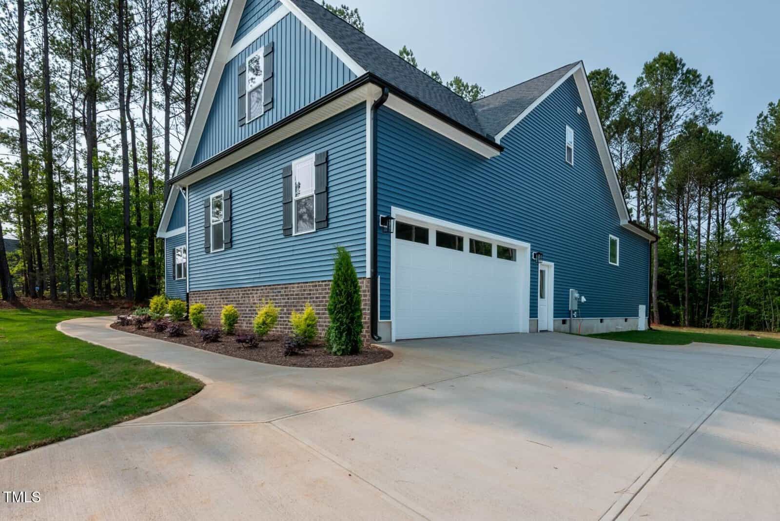 A blue house with white trim and shutters, a two-car garage, and a curved concrete driveway. There are small shrubs and mulch beds along the side of the house, and trees in the background.