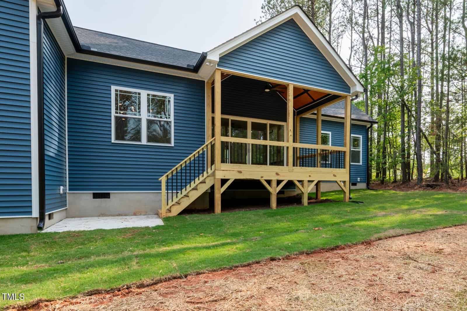 A blue house with white trim has a small covered wooden porch with stairs leading down to a grassy backyard. A wooded area is visible on the right side of the image.