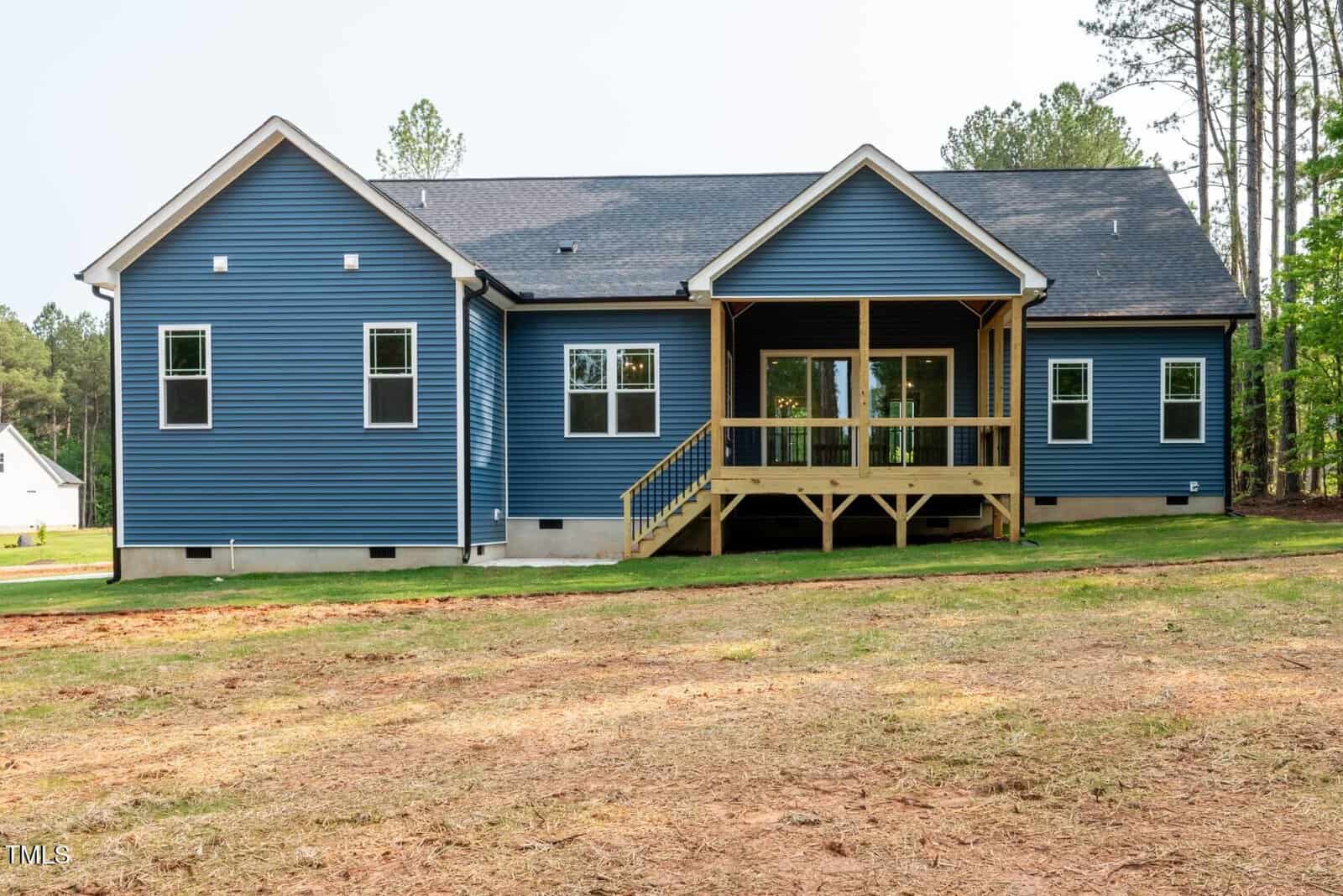 A single-story house with blue siding, white trim, multiple windows, and a small covered wooden porch at the back. The yard is mostly bare soil with some grass, and trees are visible in the background.