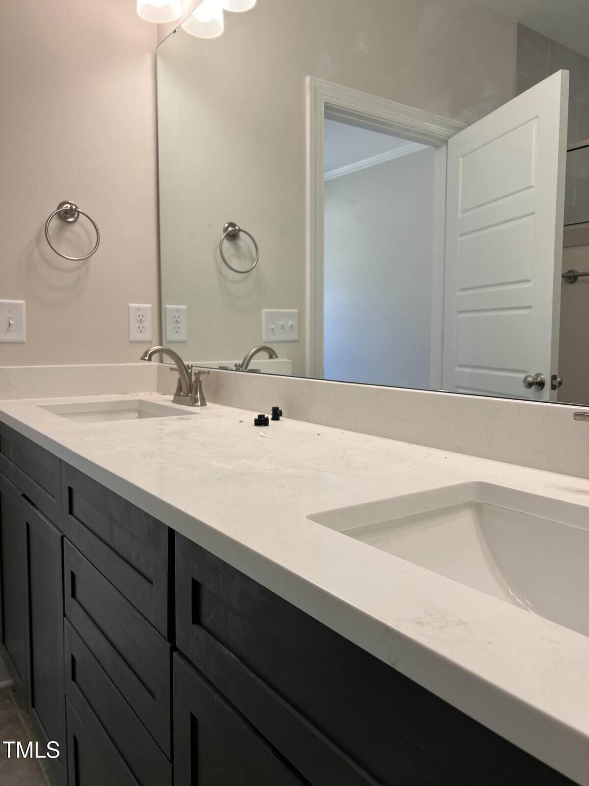 Double bathroom vanity with a white countertop, two sinks, brushed metal faucets, and a large mirror. The space includes towel rings, electrical outlets, and an open door reflected in the mirror.