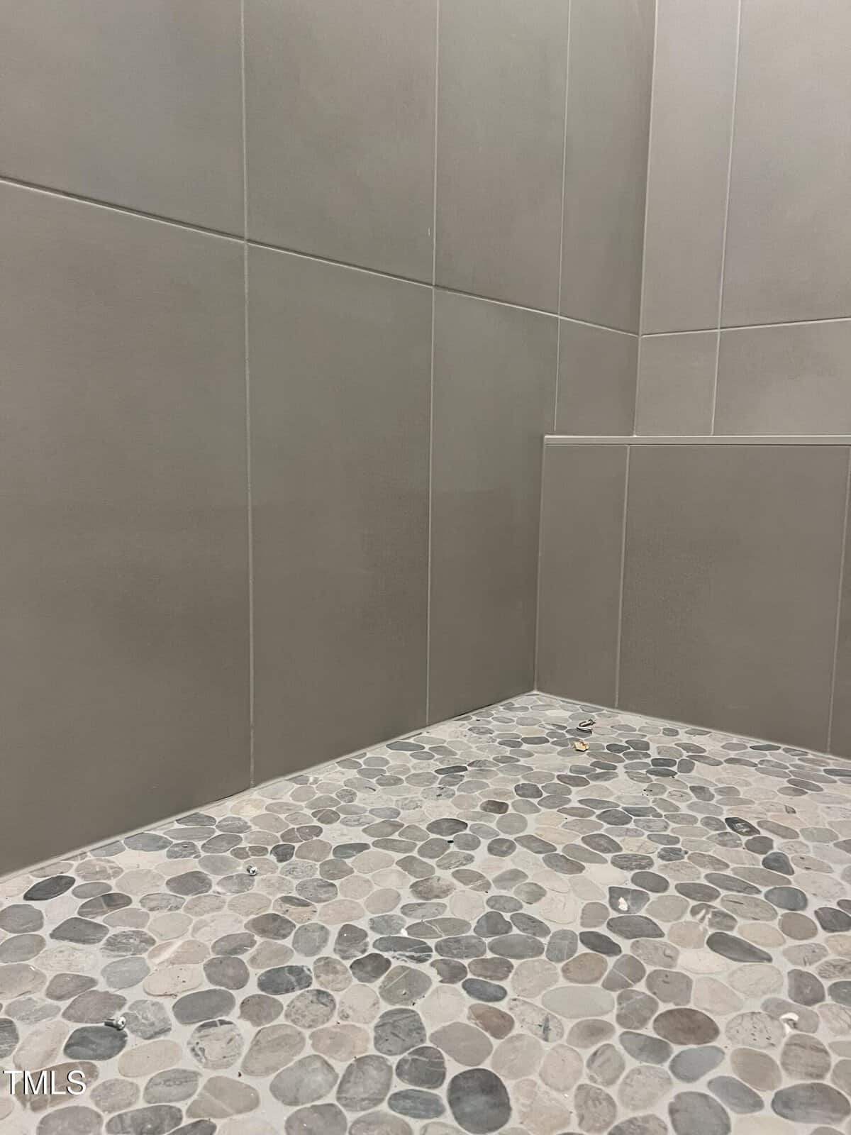 A shower corner with large beige wall tiles and a pebble-style floor made of smooth, rounded stones in various shades of gray and beige.