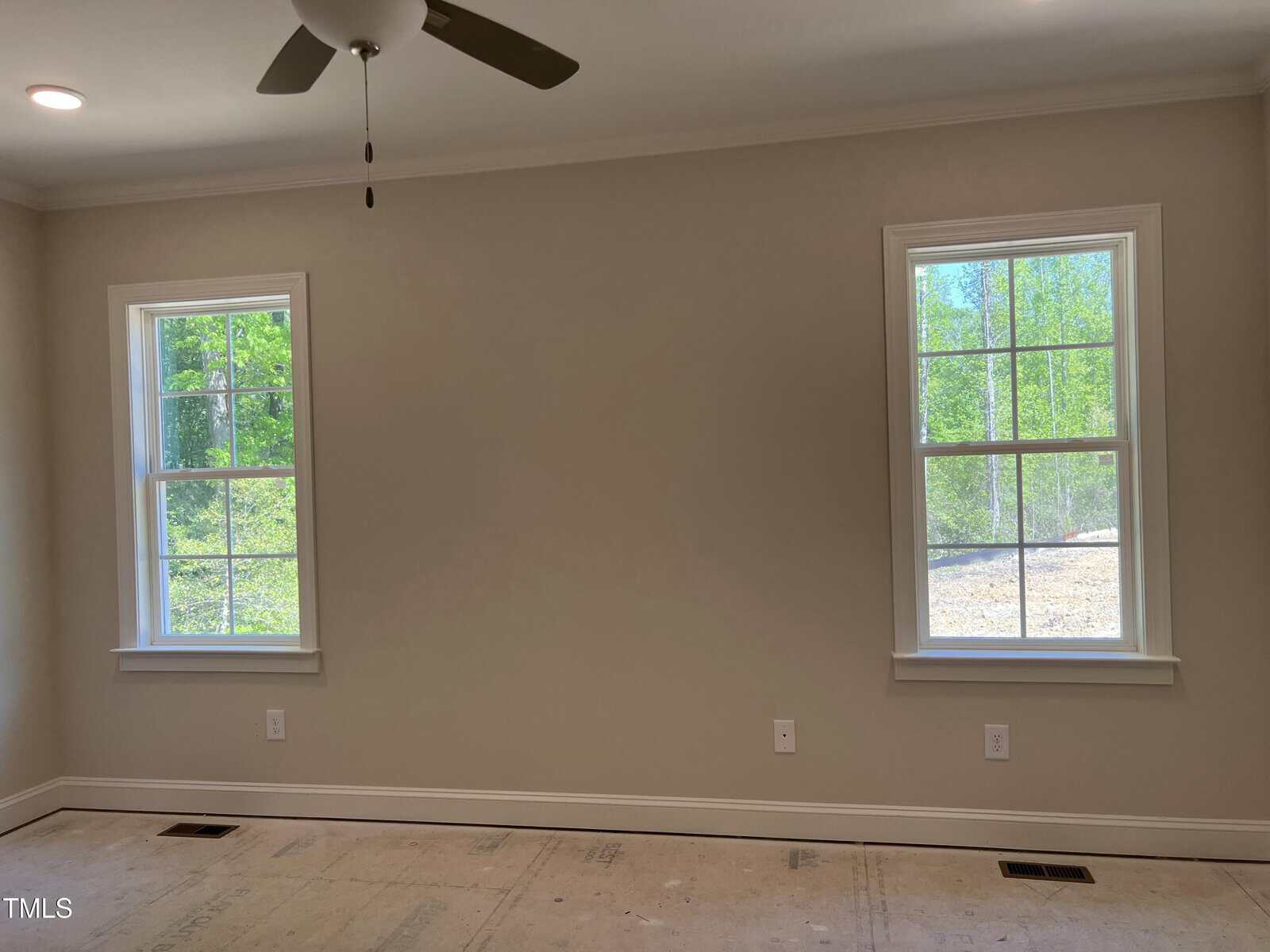 A room with light beige walls, two double-hung windows with white trim, a ceiling fan, bare floors, and visible electrical outlets below each window. The windows show green trees and natural light outside.