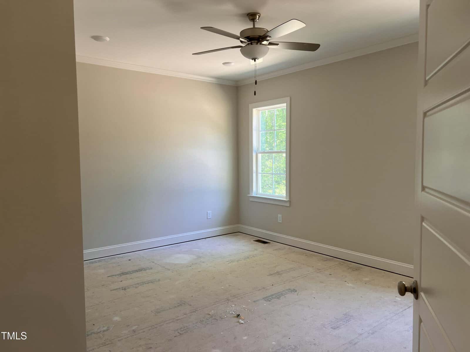 Empty room with bare floors, beige walls, white trim, a ceiling fan with light, a single window showing greenery outside, and a partially open white door with a round knob.