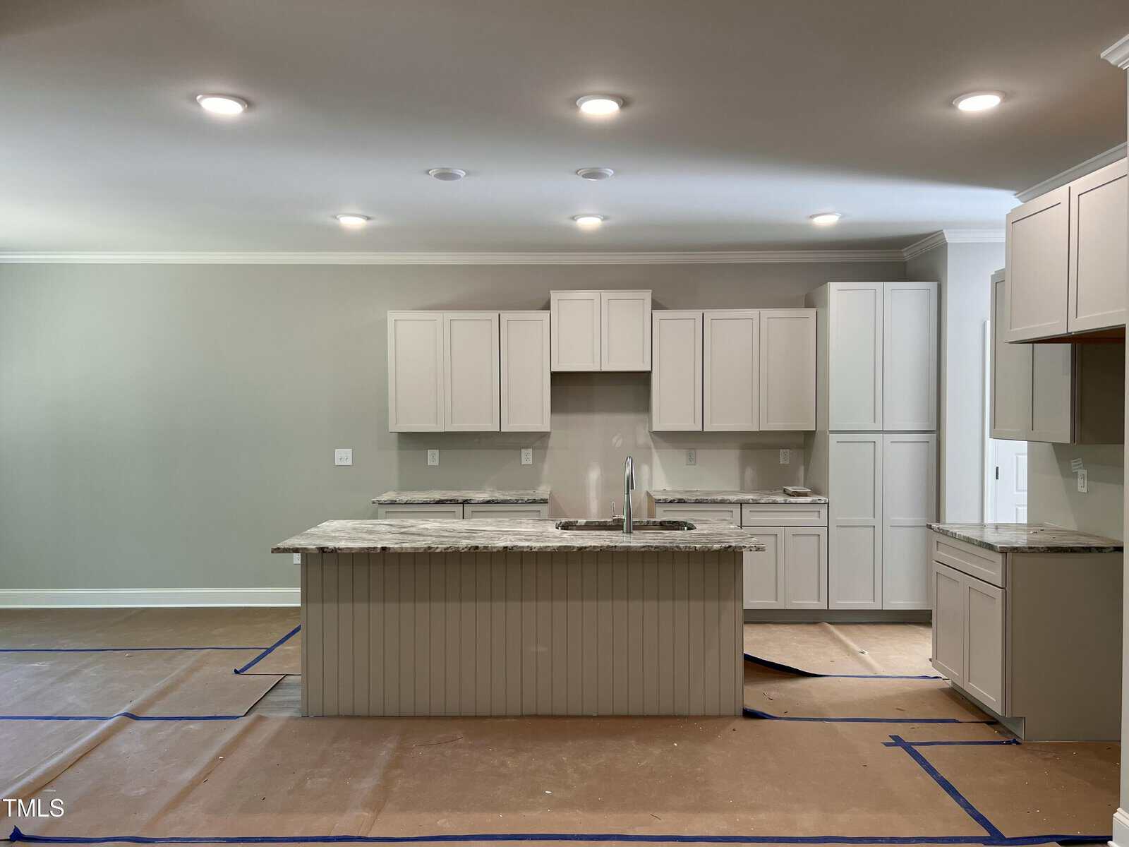 A modern kitchen with light-colored cabinets, a central island with a sink, marble countertops, recessed ceiling lights, and protective covering on the floor. Walls are a neutral color and the space appears unfinished.