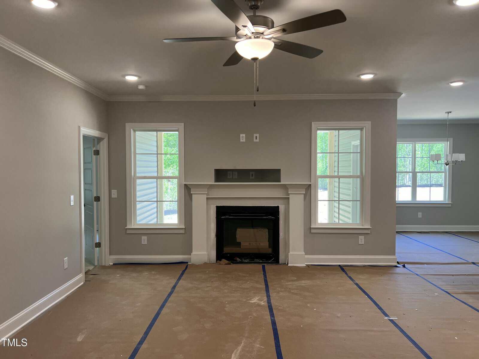 A living room under construction with cardboard and blue tape covering the floor, a ceiling fan with light, a fireplace in the center wall, and windows letting in natural light. An adjacent dining area is visible.