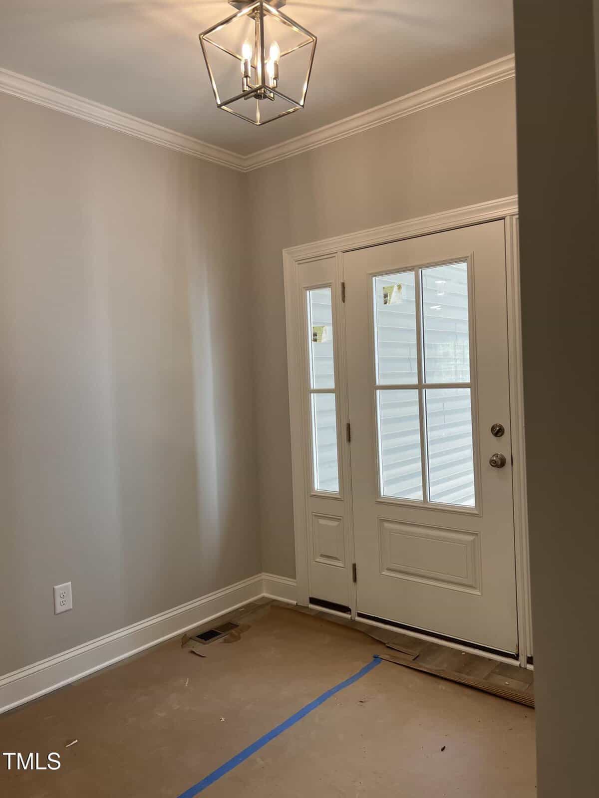 A small entryway with light gray walls, a white door with glass panels, and a modern geometric ceiling light. The floor is covered with protective cardboard and blue tape, indicating ongoing construction or renovation.