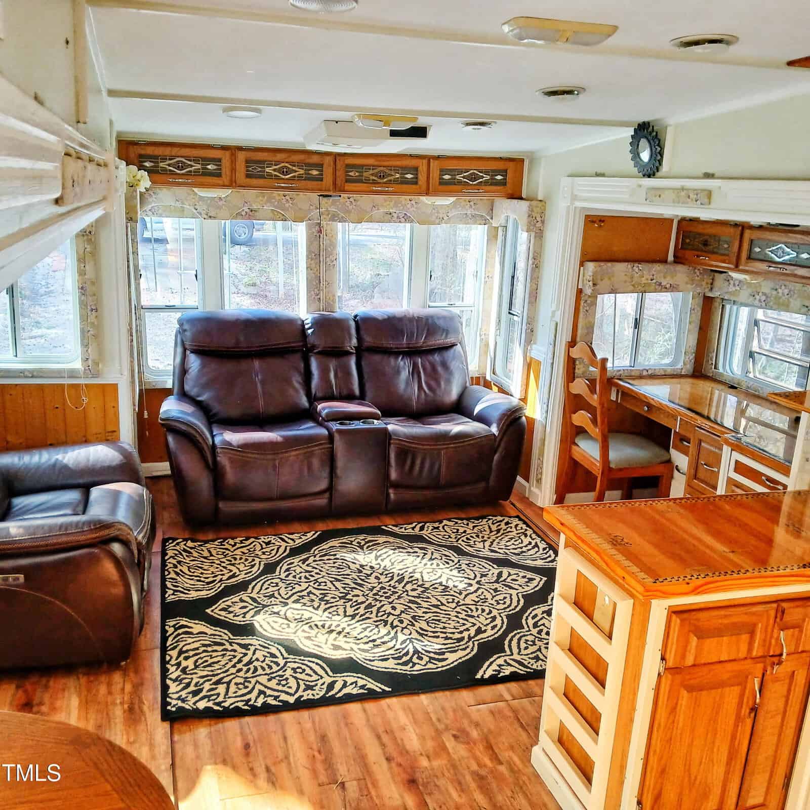 Interior view of a sunlit RV living area with brown leather seating, a patterned rug, a wood desk, and kitchen cabinetry. Large windows line the back wall, and wood accents are visible throughout.