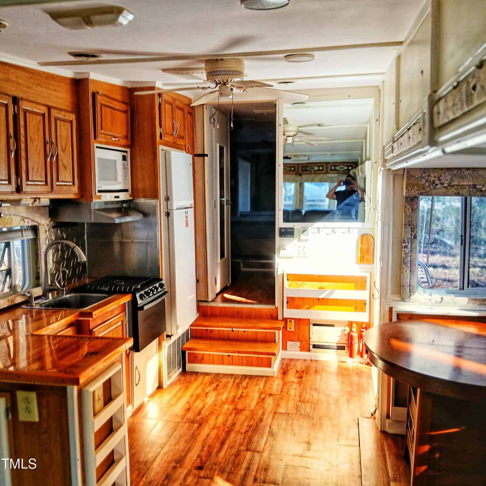 A well-lit RV interior with wood flooring, a small kitchen featuring a stove, oven, microwave, and cabinets, steps leading to a raised area, and large windows letting in natural light.