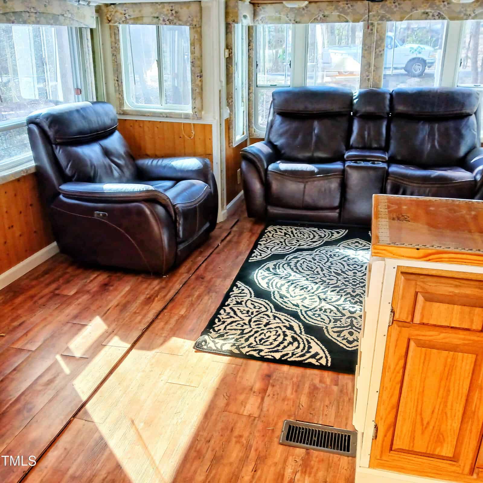 A sunlit room with wood-paneled walls and flooring, featuring a dark leather armchair, a matching leather loveseat, a patterned area rug, and part of a wooden kitchen counter in the foreground.