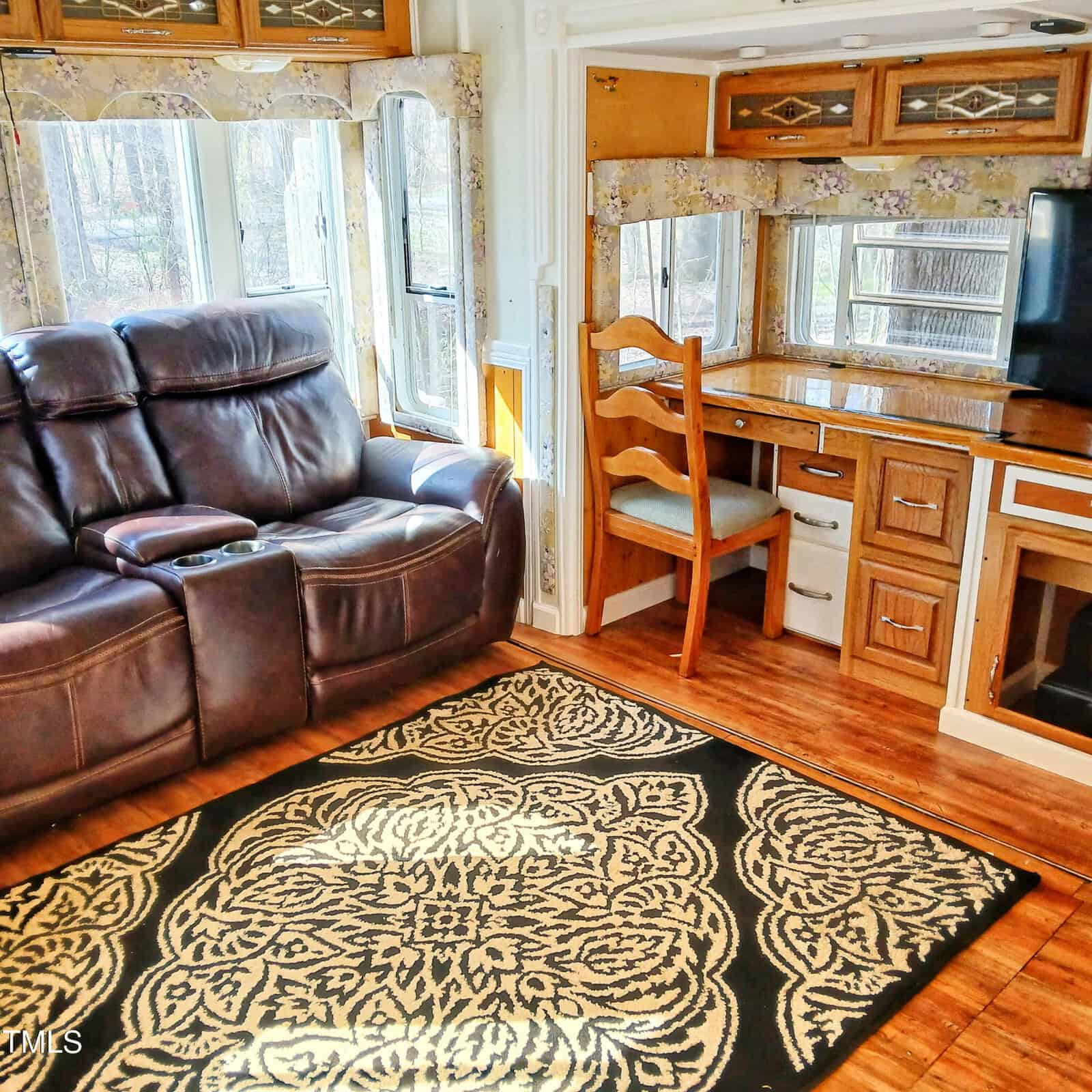 A room with a brown leather recliner sofa, a patterned rug on a wood floor, a built-in desk with a wooden chair, cabinets with glass doors, and windows letting in natural light.