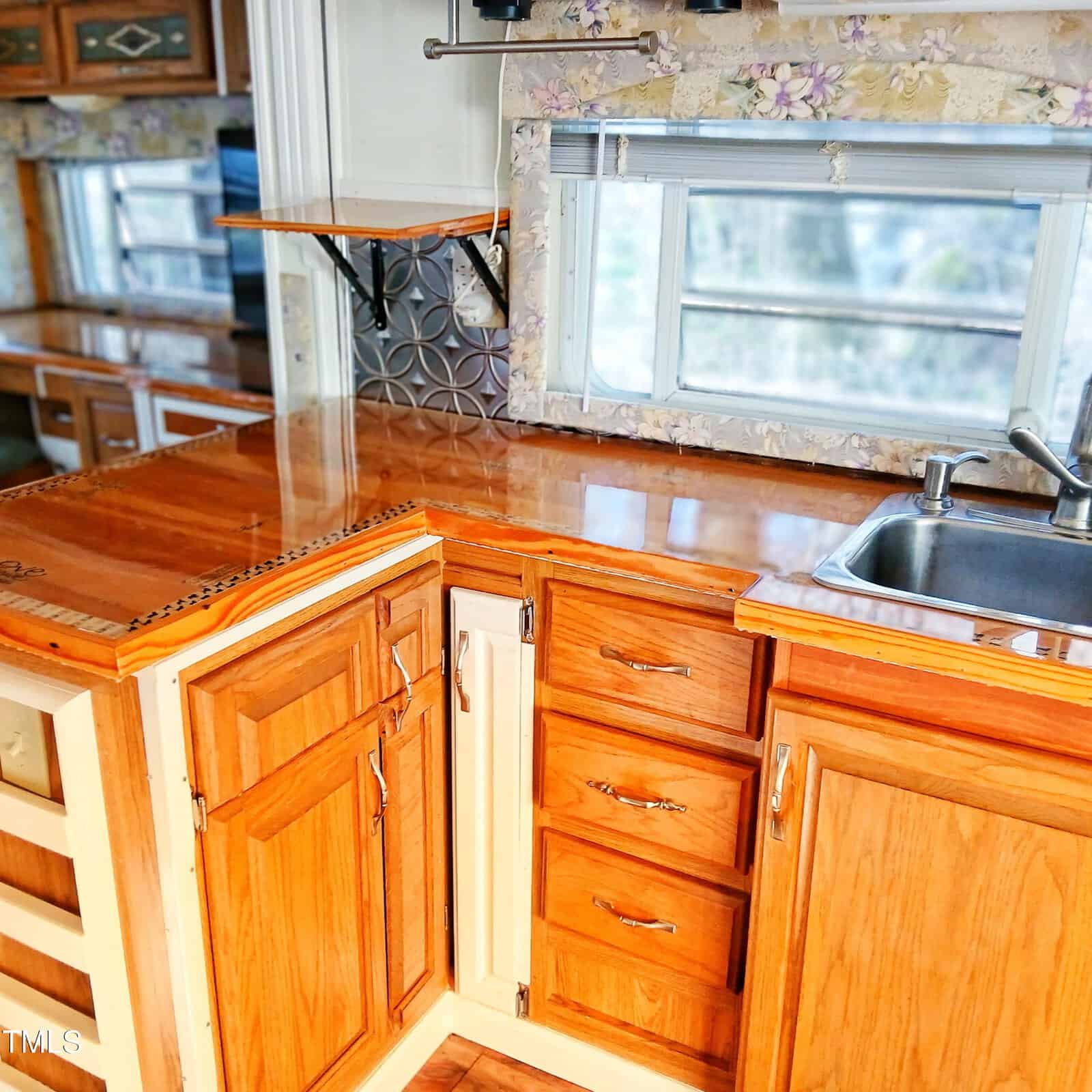 Small kitchen area with wooden cabinets, drawers, and countertops, a stainless steel sink, a window with blinds, floral wallpaper, and a tiled backsplash. Shelves are mounted on the wall above the counter.