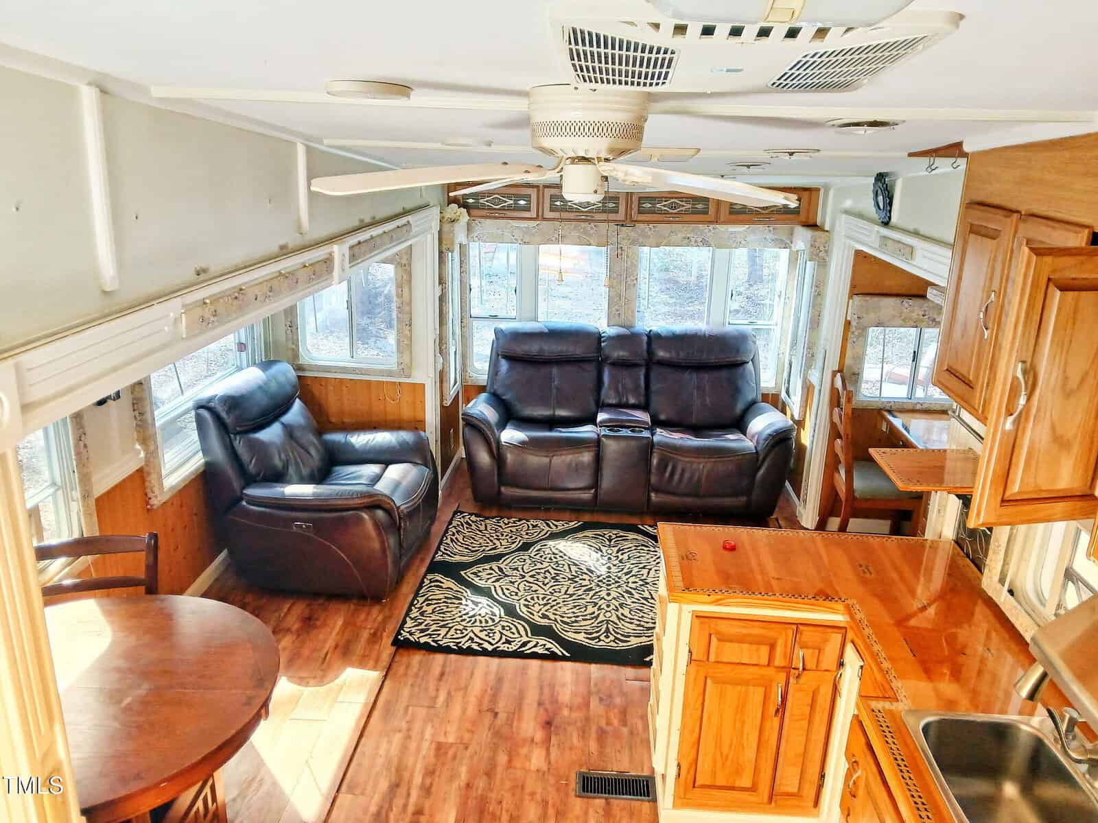 Interior of an RV featuring two leather recliners, a patterned rug, a small round dining table with chairs, wood cabinetry, a ceiling fan, and large windows letting in natural light.