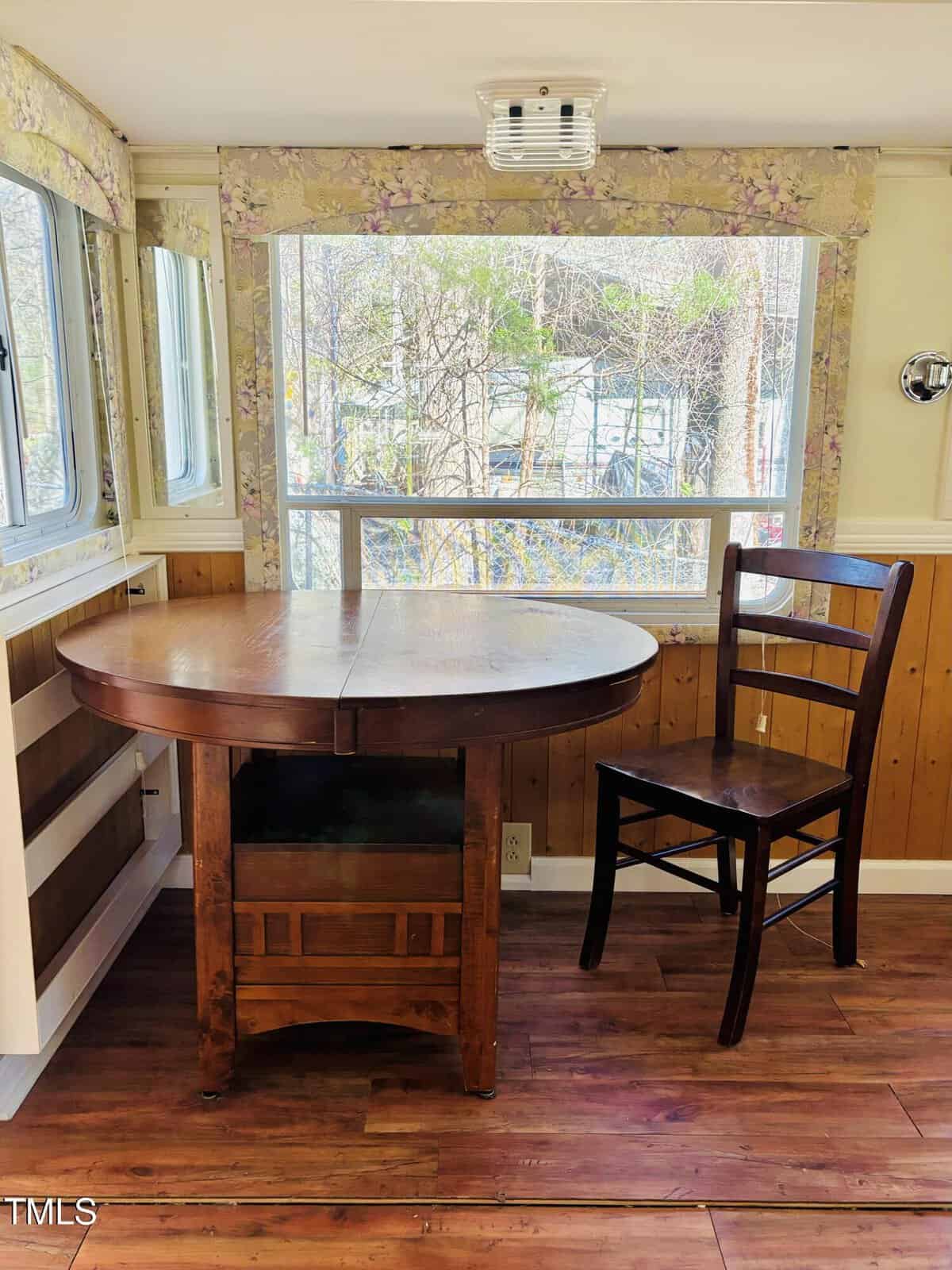A wooden table with drop leaves and a wooden chair are placed in front of a large window with floral curtains. Sunlight comes through the window, and trees are visible outside. The floor and lower walls are wood-paneled.