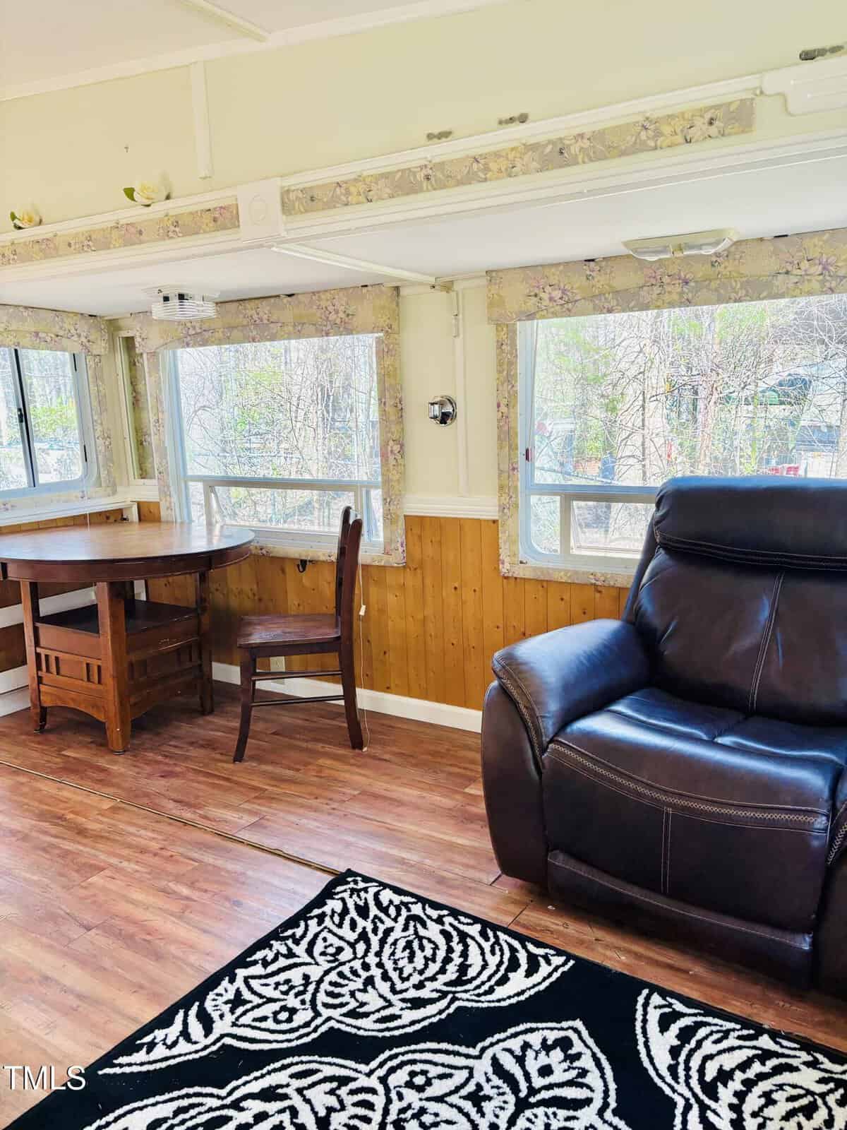 A room with wood-paneled walls, floral curtains, large windows, a round wooden table with a chair, a black leather recliner, and a black-and-white patterned rug on a wood floor.