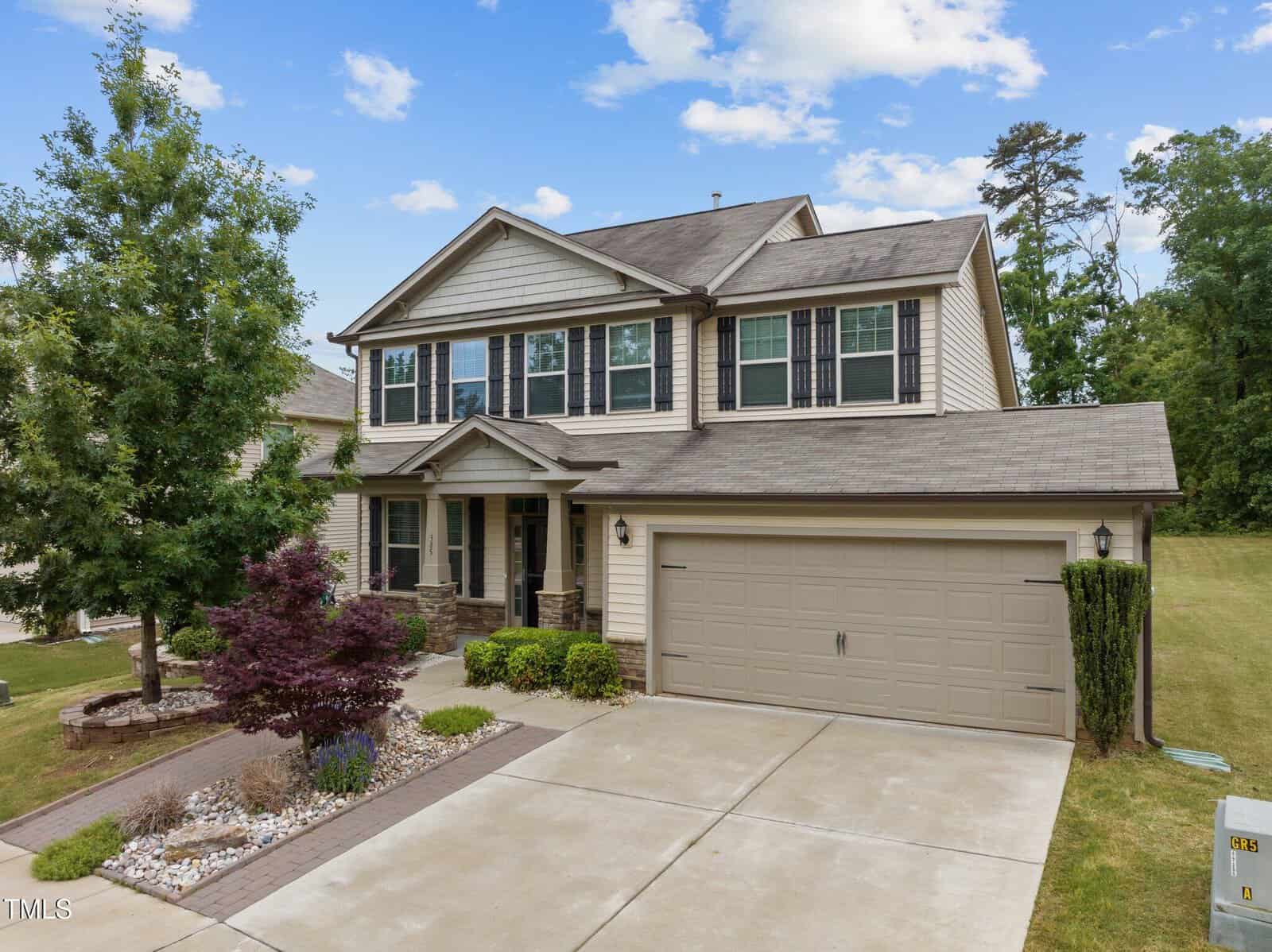 Two-story suburban house with beige siding, dark shutters, and a double garage. The front yard has a small landscaped area with shrubs, a tree, and decorative stones. A concrete driveway leads to the garage.