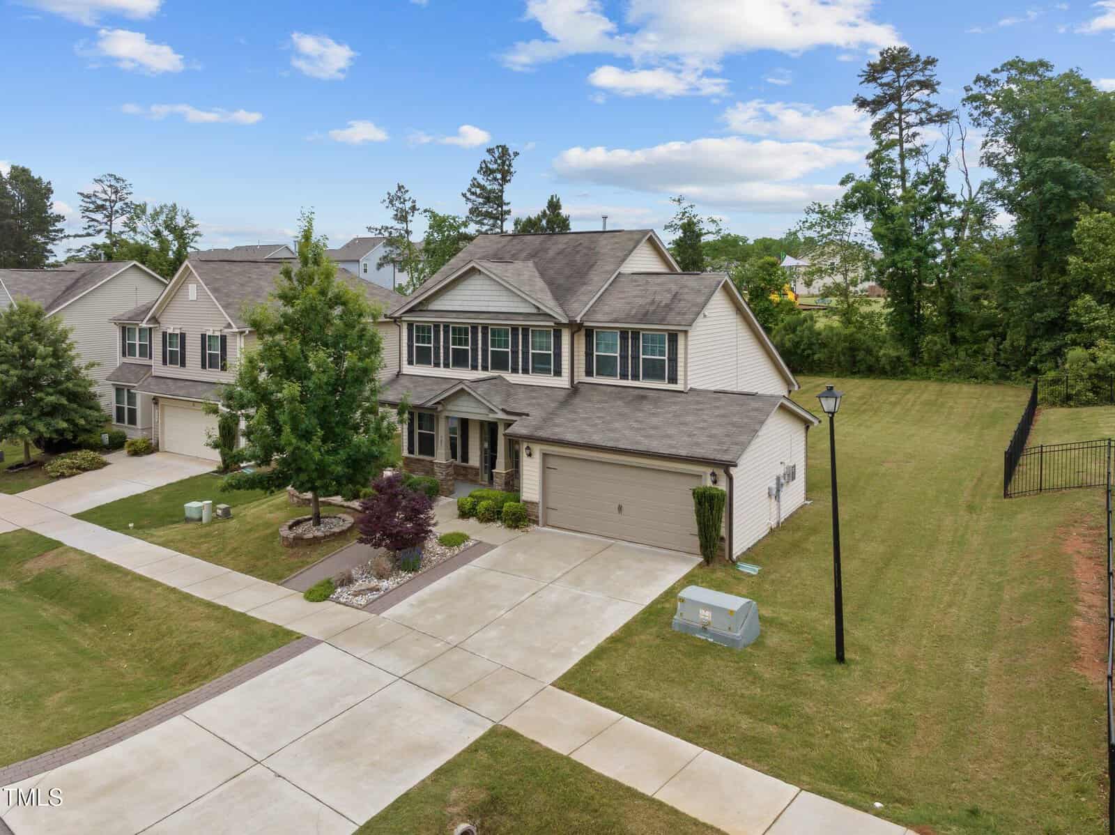 A two-story suburban house with gray siding, a double garage, and a well-maintained lawn. The home is surrounded by similar houses and greenery, with a clear sky and some scattered clouds overhead.