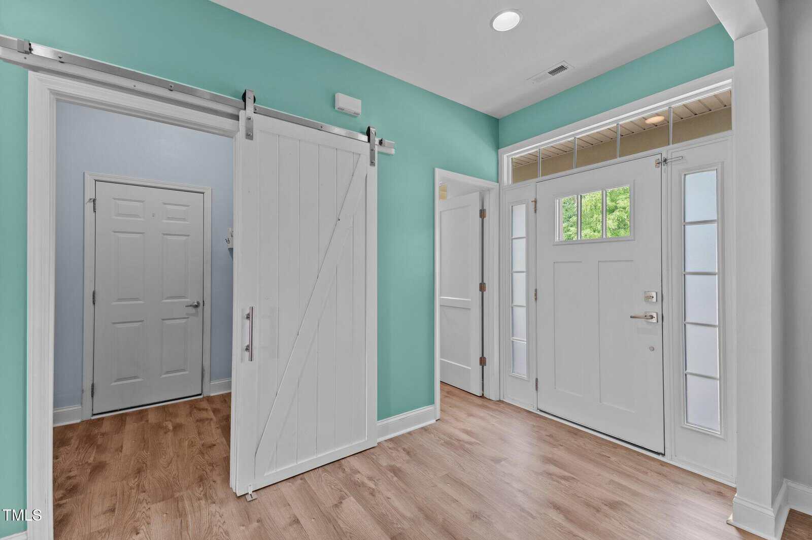 A bright entryway with a white front door featuring glass panels, sidelights, and transom windows. The space has teal walls, wood flooring, and a white barn door leading to an adjacent room.