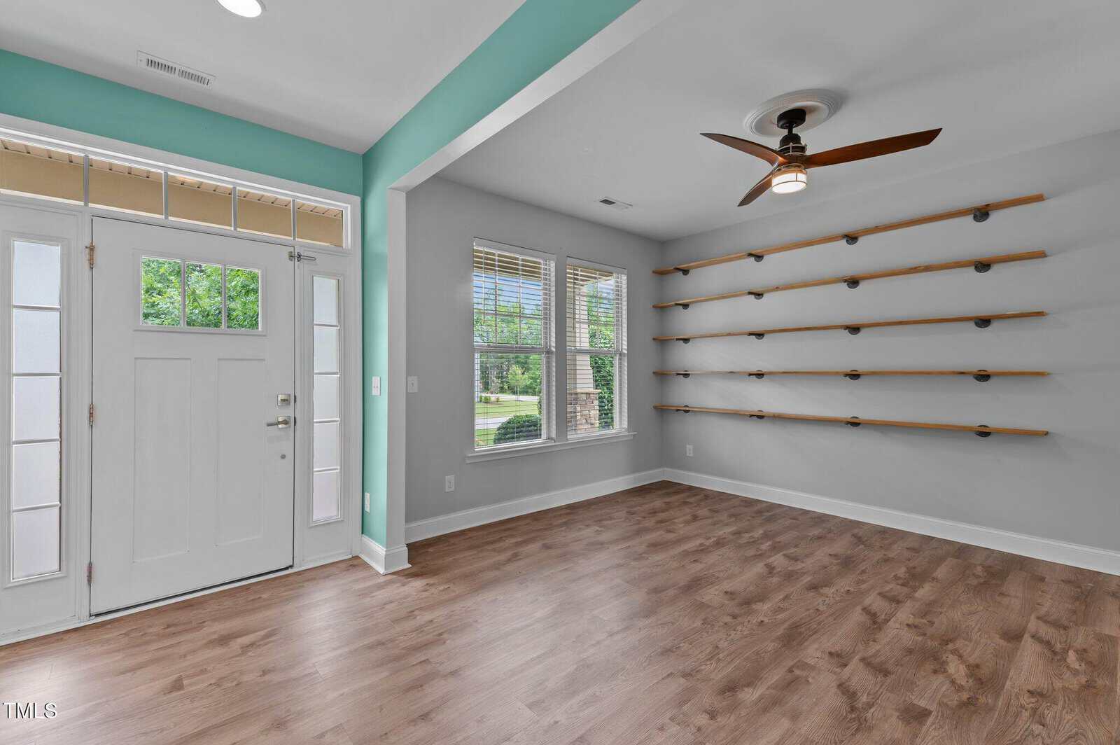 A bright room with wood flooring, a ceiling fan, a teal accent wall, a white front door with sidelights, three windows, and a wall-mounted shelving unit with wooden shelves and black brackets.