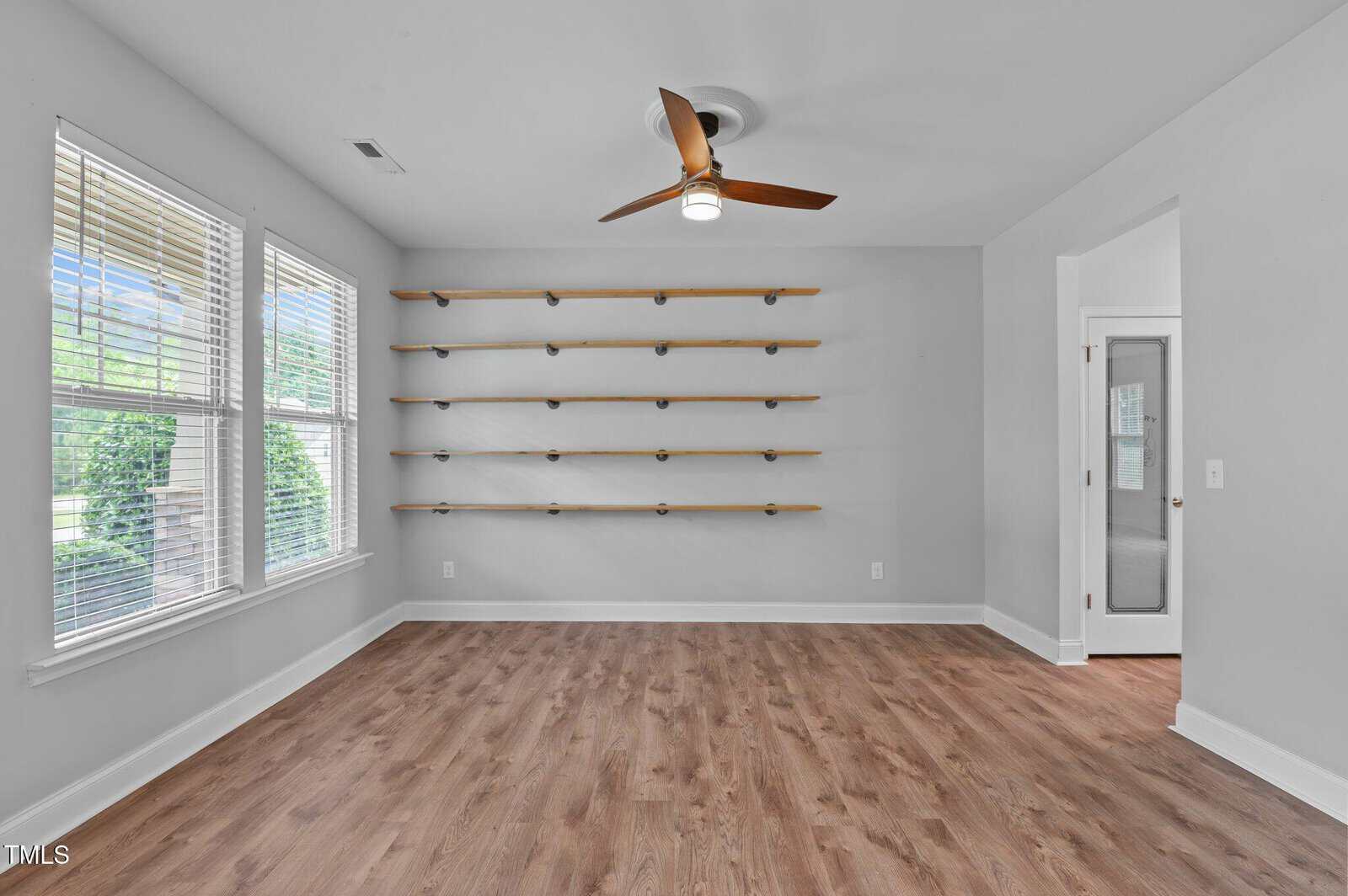 A room with wood-look flooring, a ceiling fan, a wall with six wooden shelves, two large windows with blinds on the left, and a door with frosted glass on the right. The walls are painted light gray.