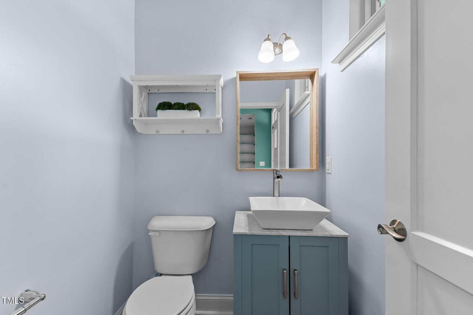 Small bathroom with light blue walls, a white toilet, a blue vanity with a square vessel sink, a wall-mounted mirror, and a white shelf with a plant. The door is open, showing another room in the background.