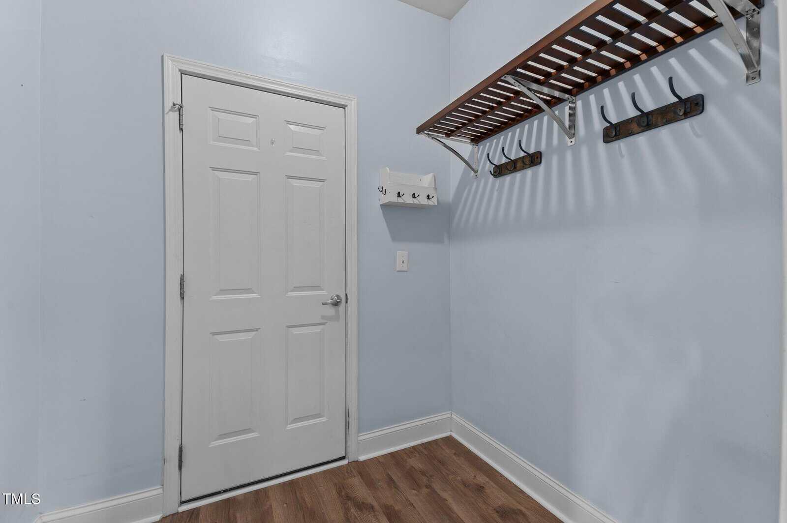A small room with light blue walls, a white door, wood flooring, a wooden shelf with hooks on the right wall, and a smaller white hook rack next to the door.
