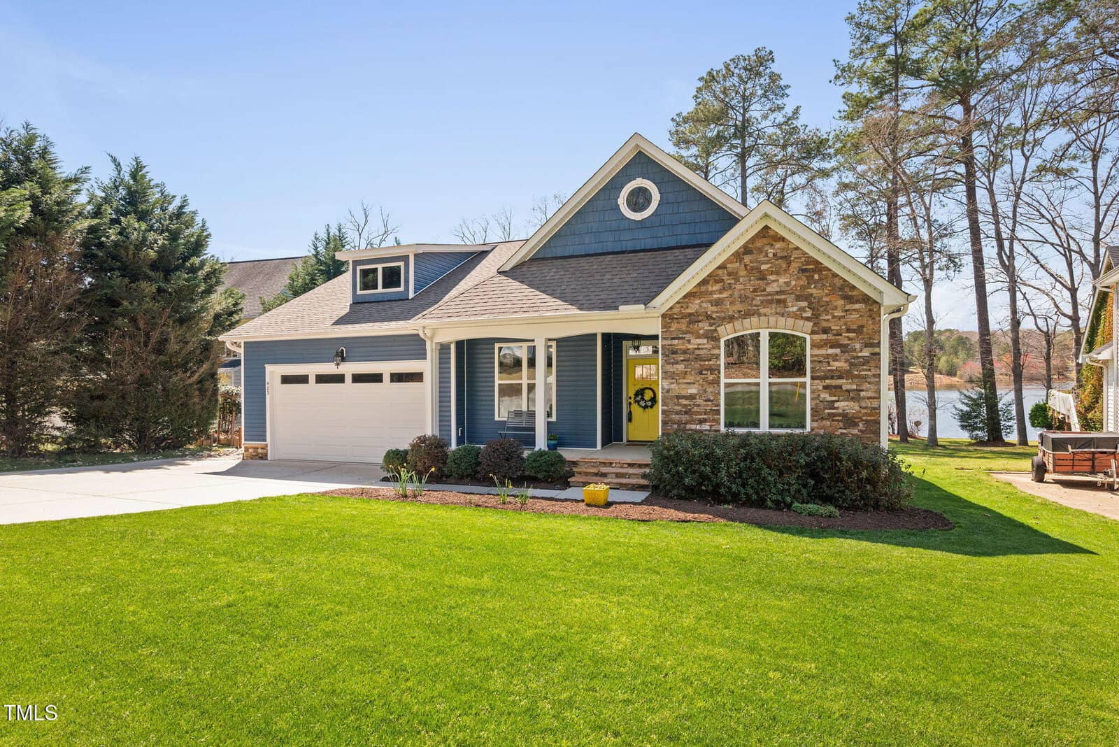 A single-story house with blue siding, stone accents, white trim, a covered front porch, two-car garage, and a well-maintained green lawn. Trees are in the background, and a lake is visible to the right.
