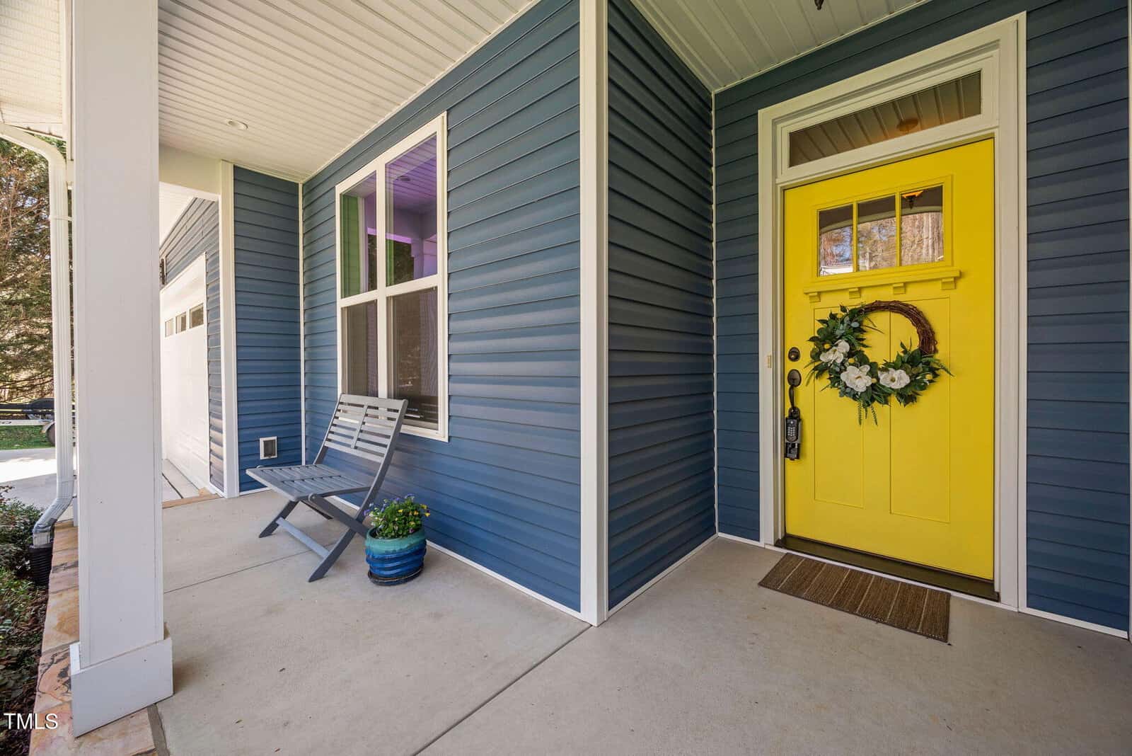 A small front porch with blue siding, a bench, a potted plant, and a bright yellow front door decorated with a floral wreath. A brown doormat lies in front of the door.