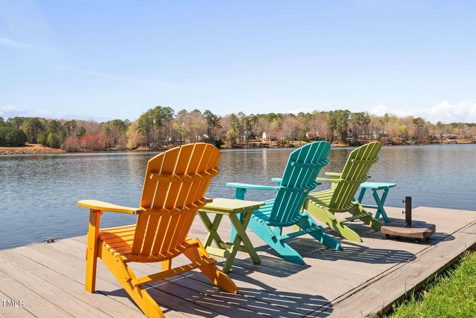 Three colorful Adirondack chairs—yellow, orange, and green—sit on a wooden dock facing a calm lake with trees and houses visible across the water under a clear blue sky.