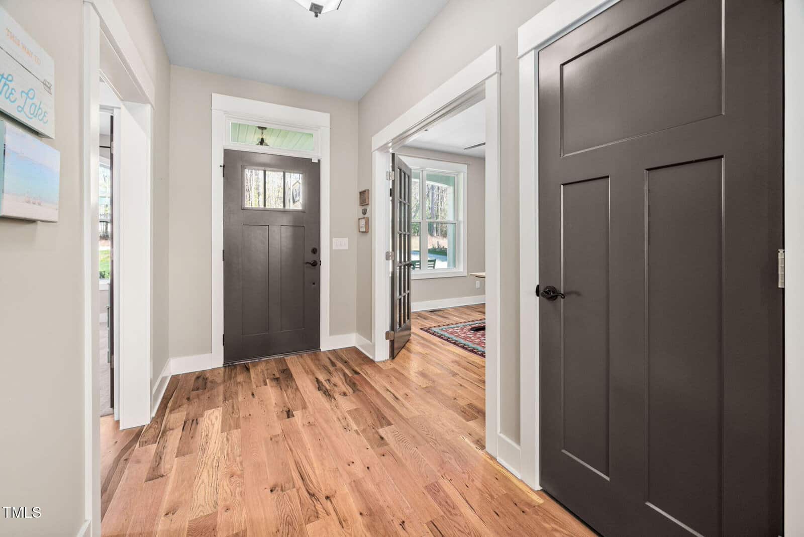 A hallway with light-colored walls, wood flooring, and dark brown doors. Natural light comes through windows in adjacent rooms. The entry door is centered, and white trim frames doorways and windows.