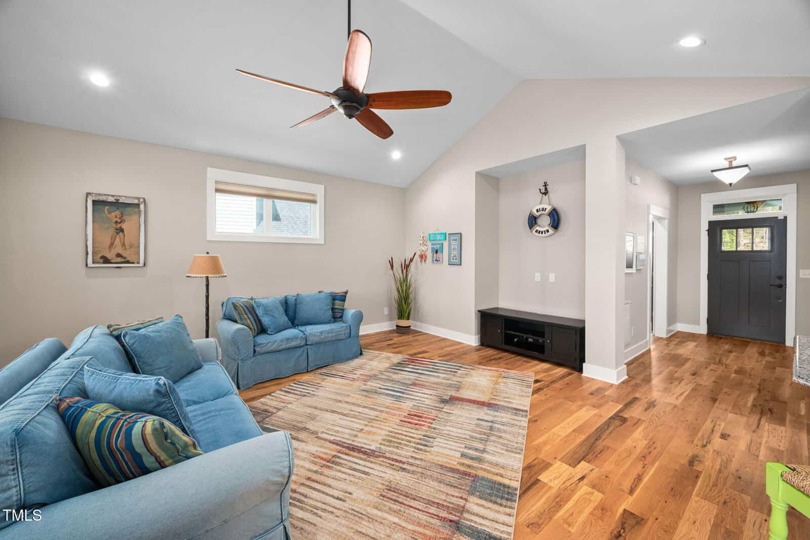 A living room with wood flooring, blue sofas, a colorful striped rug, a ceiling fan, and a built-in entertainment nook. The walls are light beige, and the front door and hallway are visible in the background.