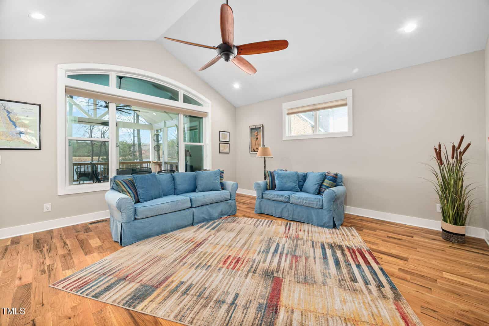 Living room with two blue sofas, a multicolored area rug, hardwood floors, and a ceiling fan. Large windows let in natural light, and potted plants and framed pictures decorate the neutral-colored walls.