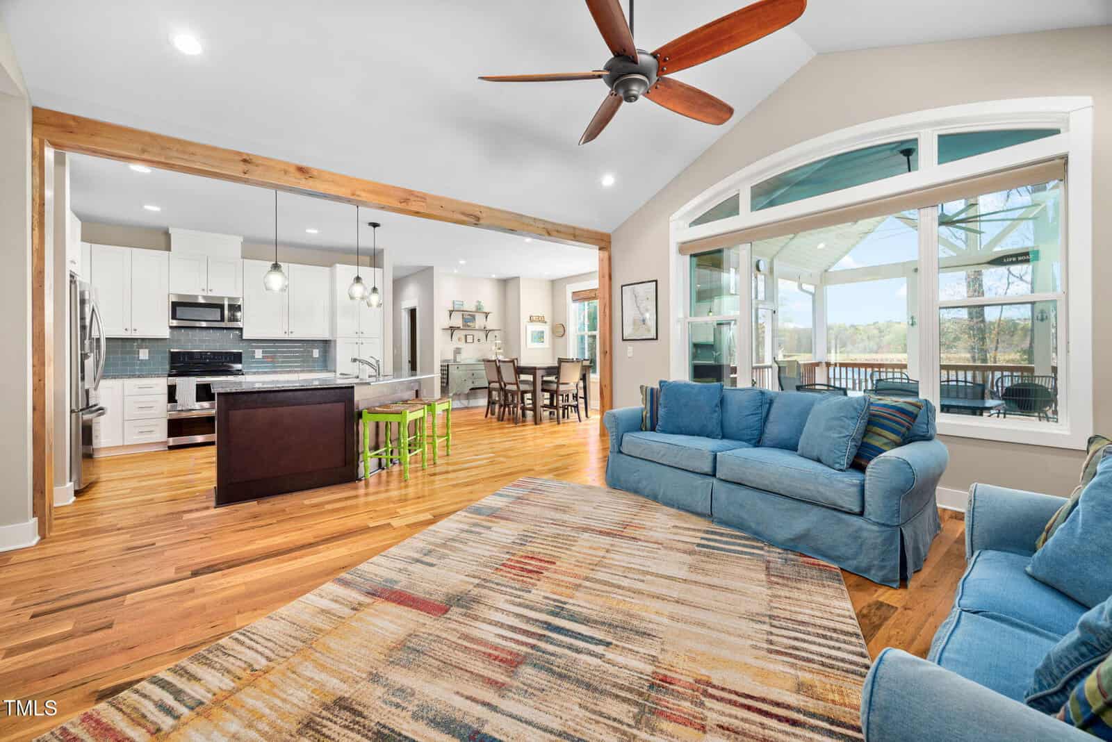 Open living area with blue sofas, a multicolored rug, hardwood floors, and ceiling fan. The space connects to a kitchen with an island, green stools, and pendant lights, and a dining area near large windows and a patio.