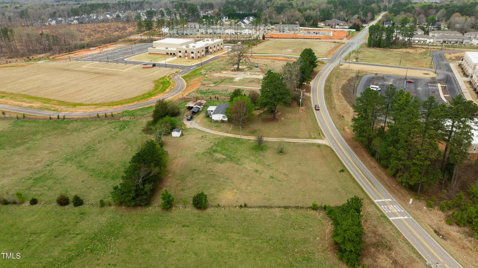 Aerial view of a rural area with an open field, a few buildings and trees, a two-lane road curving through the landscape, and a large school building with sports fields in the background.