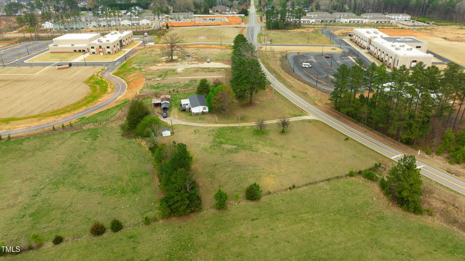 Aerial view of a rural area with open grassy fields, a few scattered trees, two white buildings near the center, a curved road, and larger commercial buildings on either side of the road in the background.